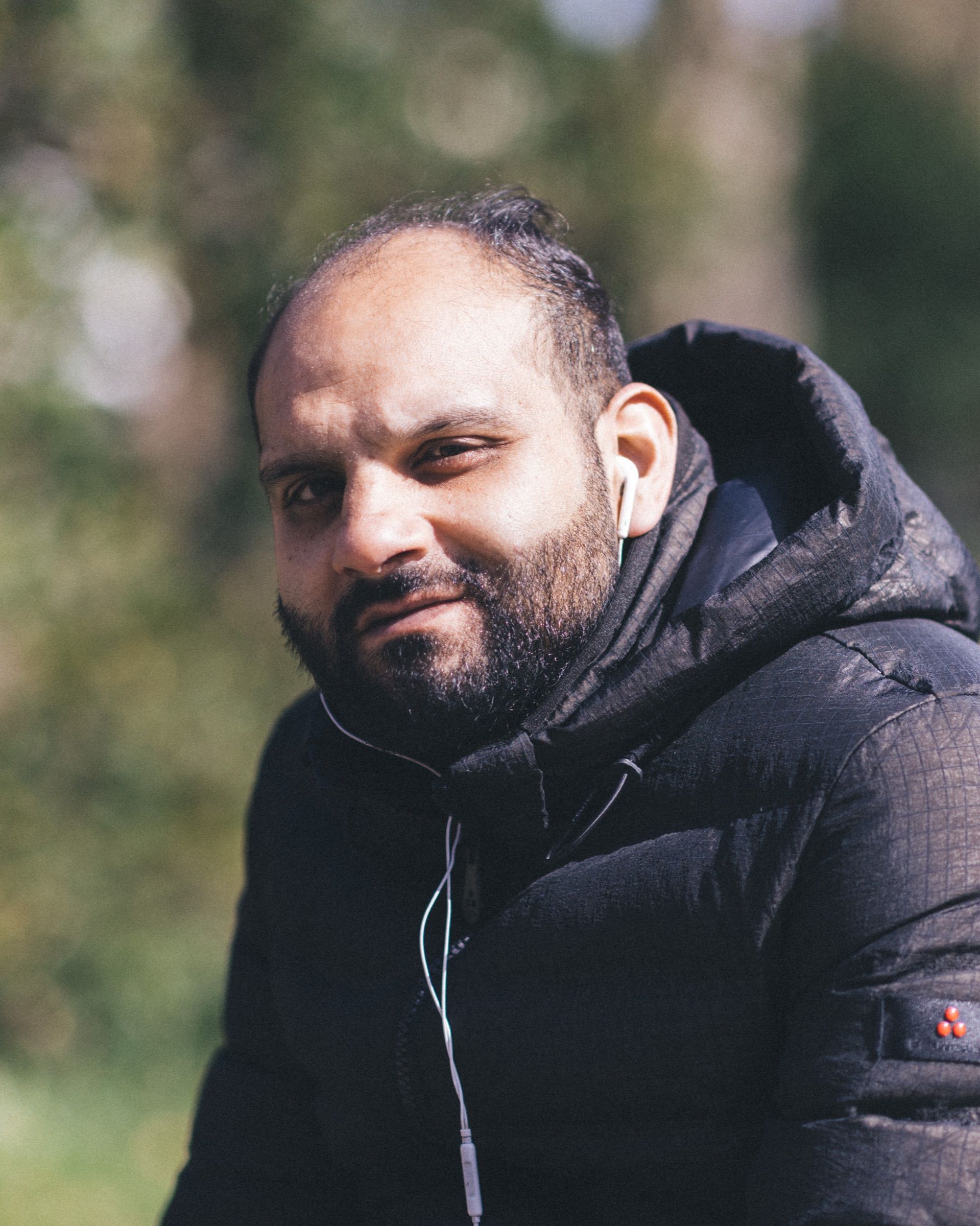 A man in a black puffer jacket, earphone cable hanging, looks into the camera with sunlit trees behind him.