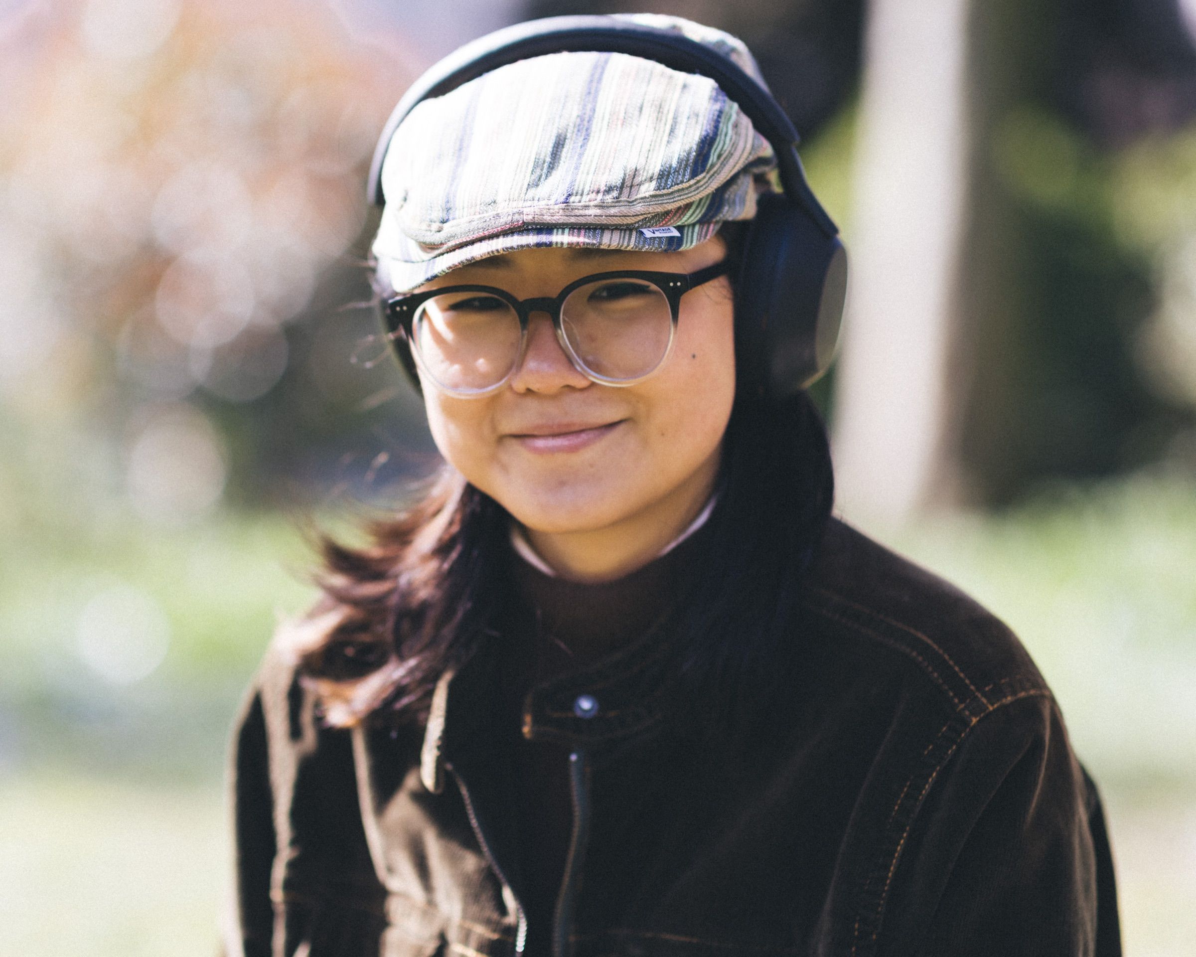 A woman in a flat cap, round glasses and headphones smiles in spring sunlight.