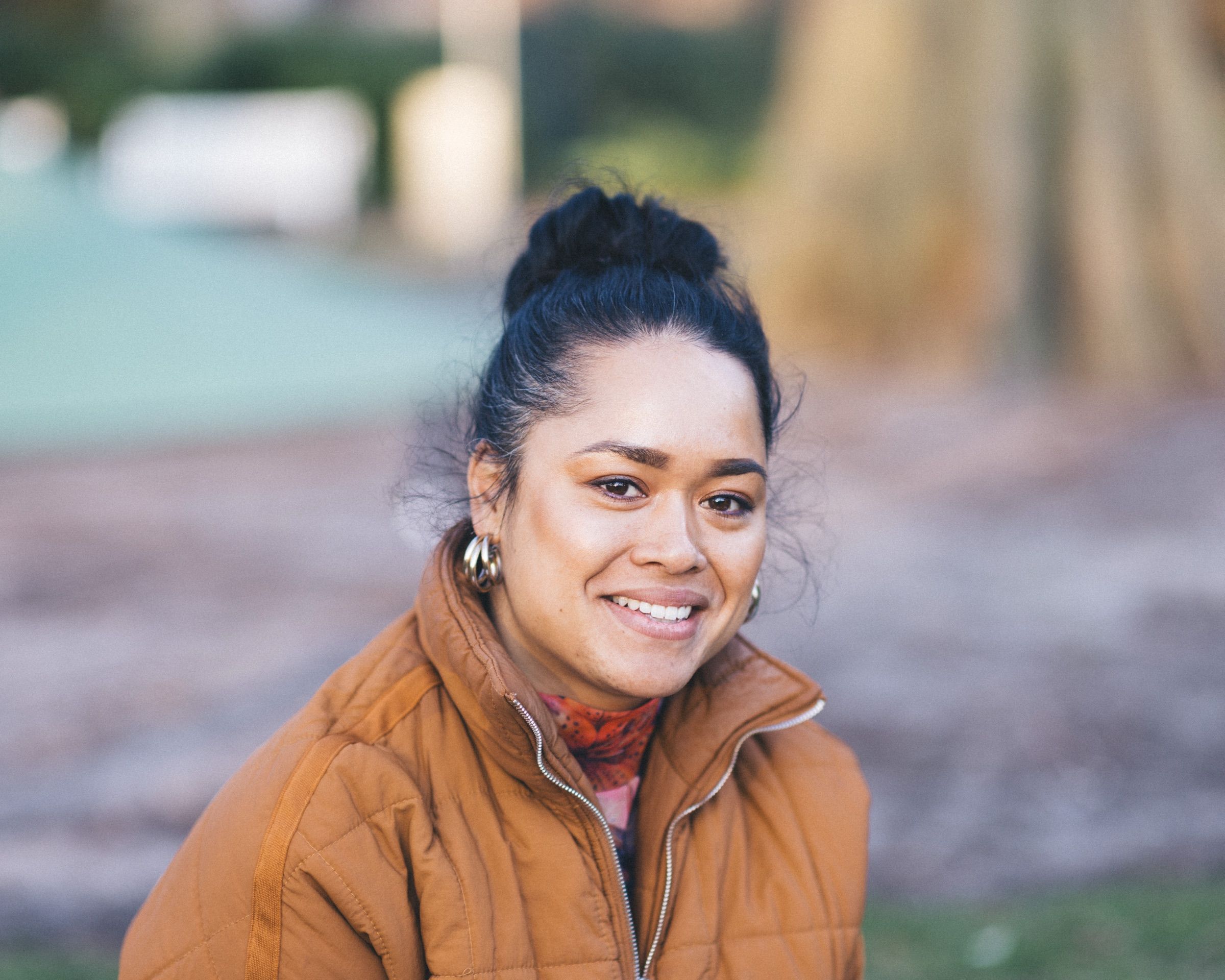 A woman with her hair up, in an ochre jacket, smiles with the park soft behind her.