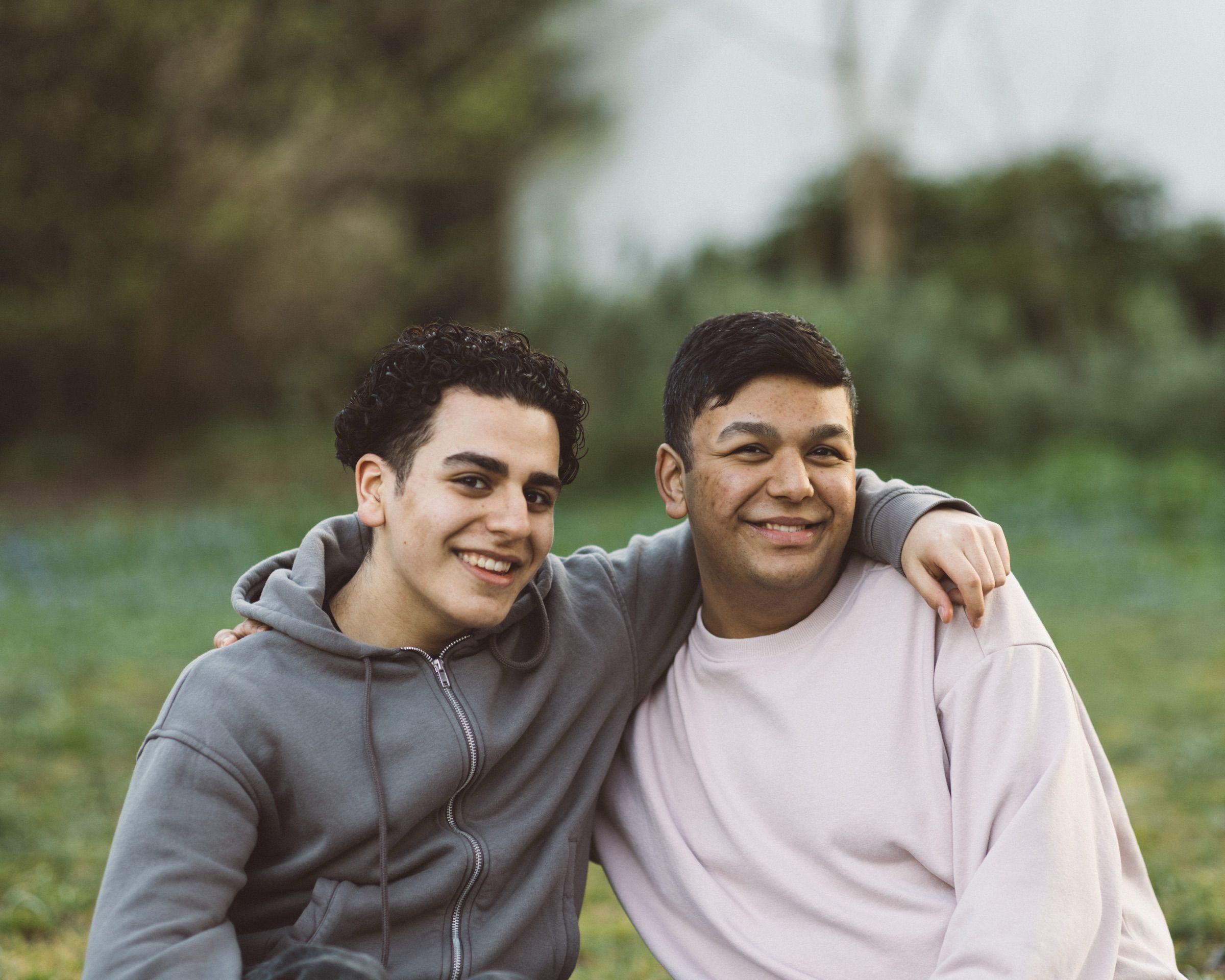 Two young men in hoodies stand arm in arm on a lawn, grinning.