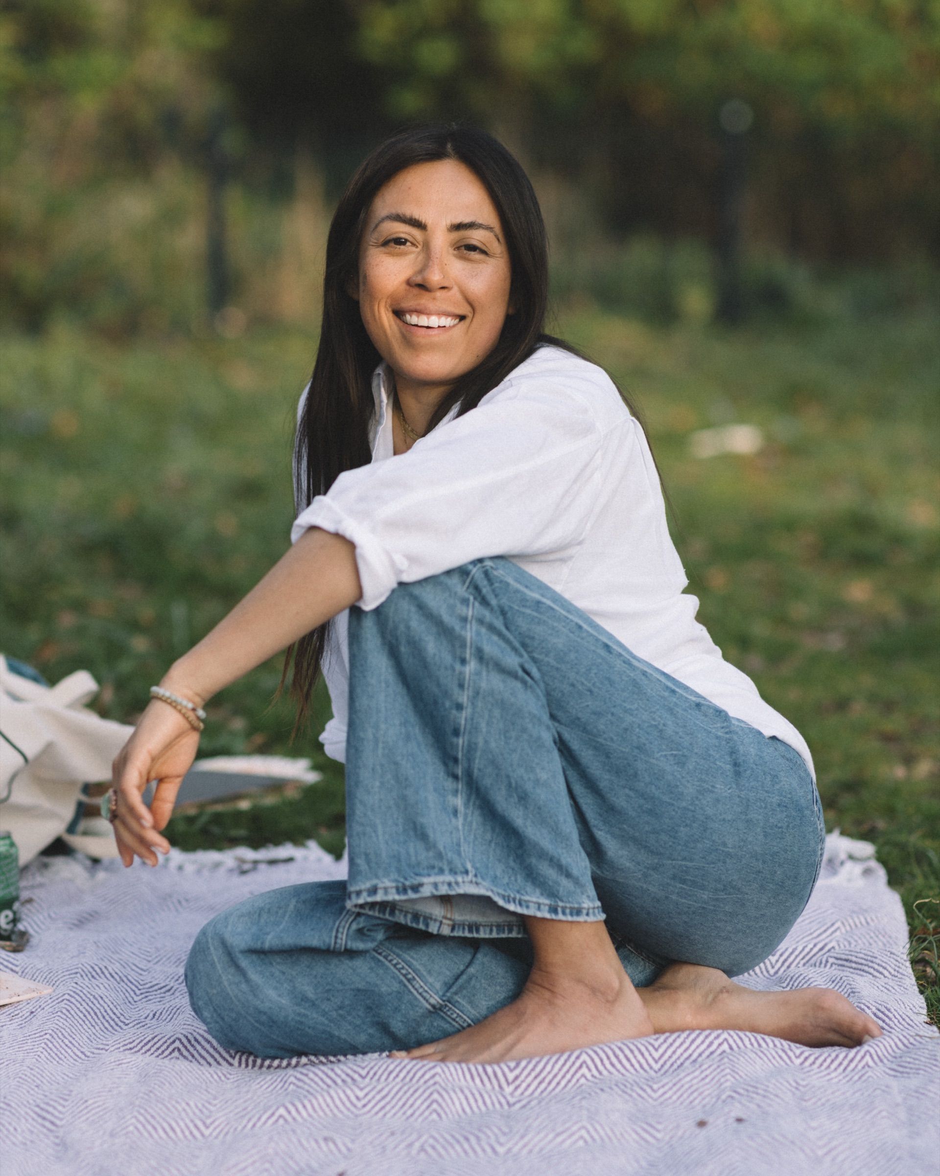 A woman in a white shirt and jeans sits barefoot on a blanket in the grass, smiling.