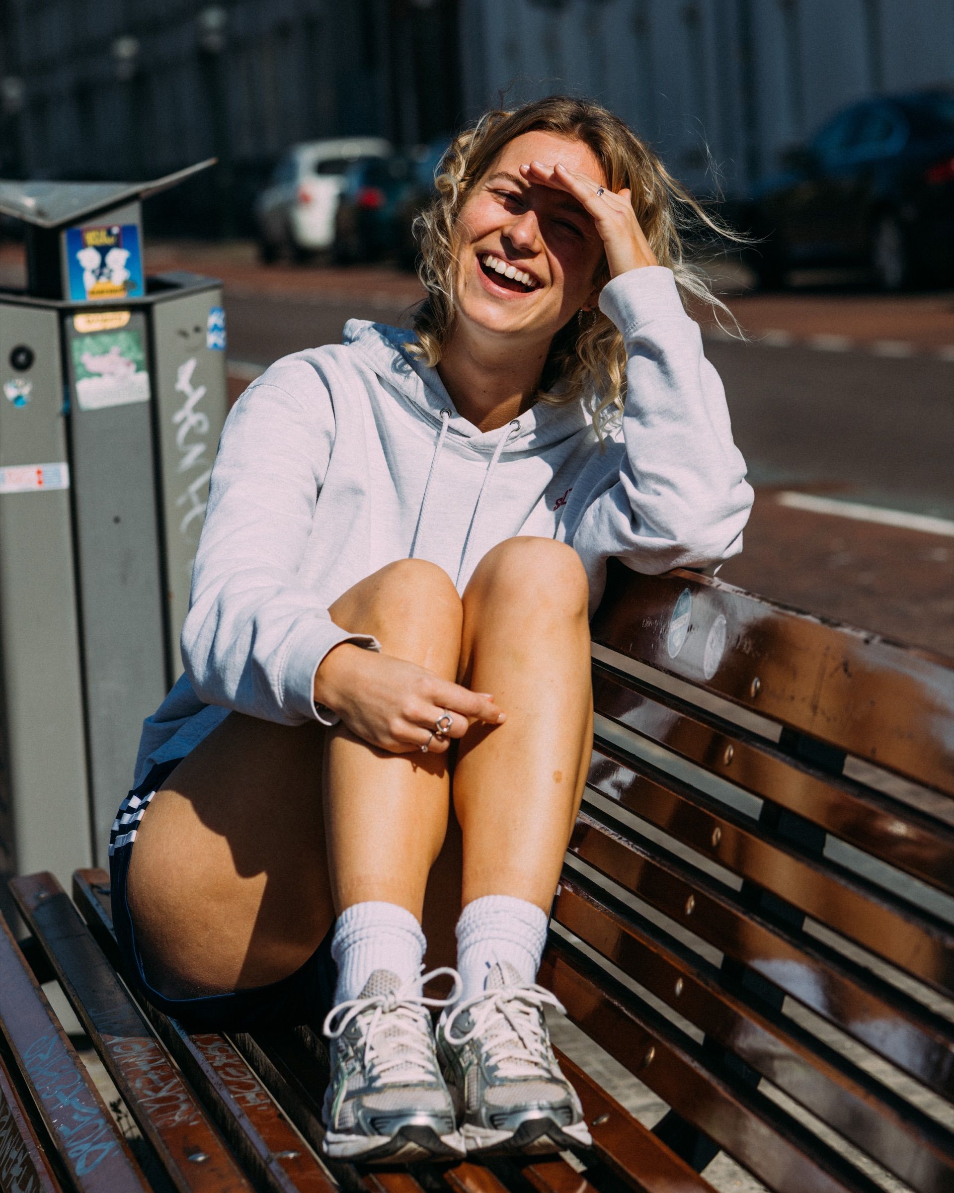 A woman in a grey hoodie laughs on a wooden bench by the street, one hand in her hair.