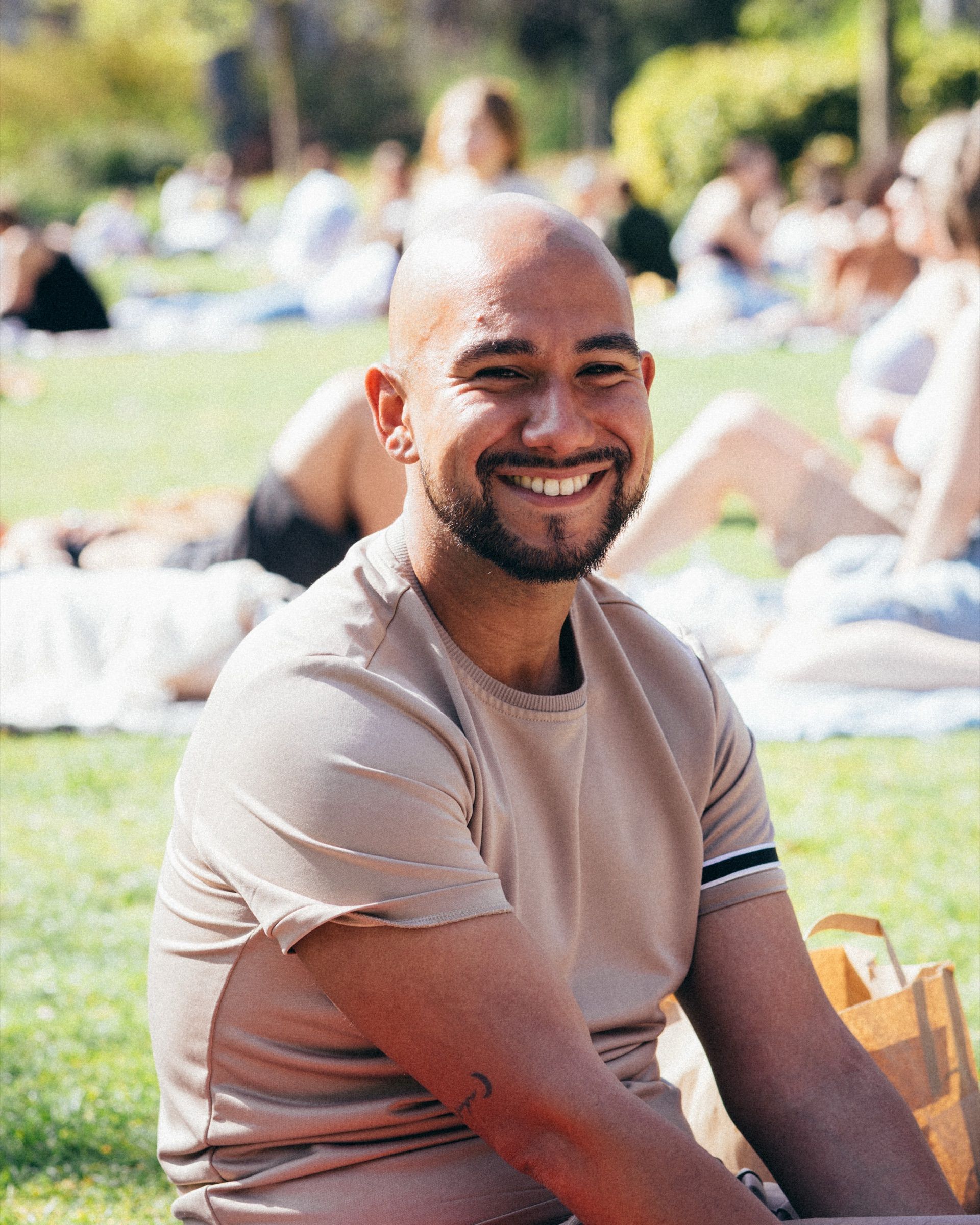 A man in a beige T-shirt smiles broadly, a crowded summer lawn behind him.