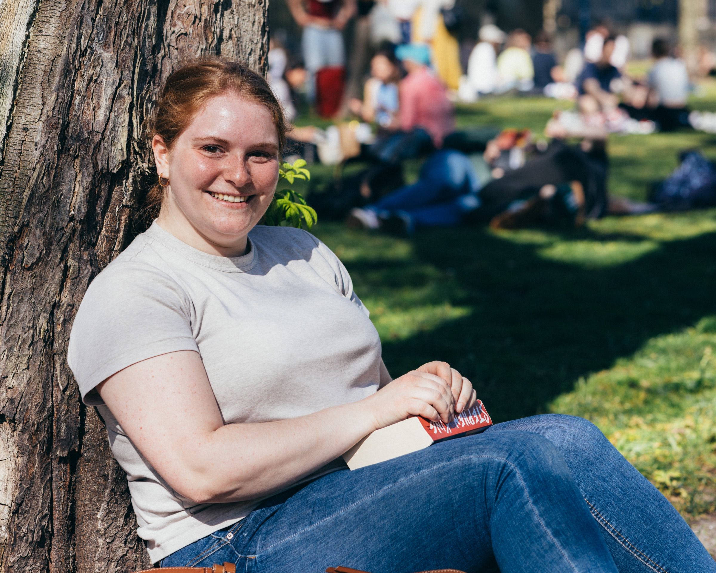A woman in a grey T-shirt rests against a tree on a busy lawn, smiling.