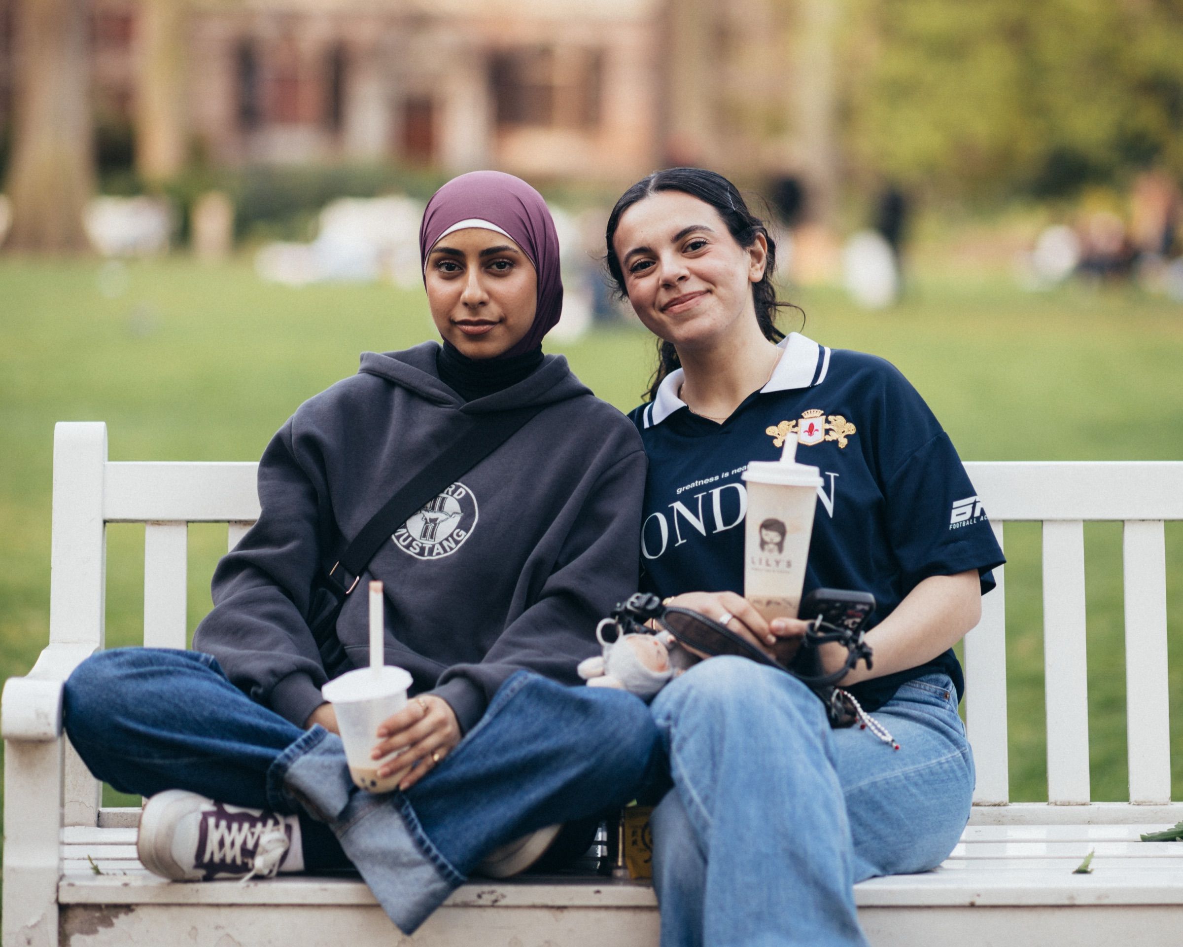 Two friends sit on a white bench with drinks in their hands, one in a headscarf, both smiling.