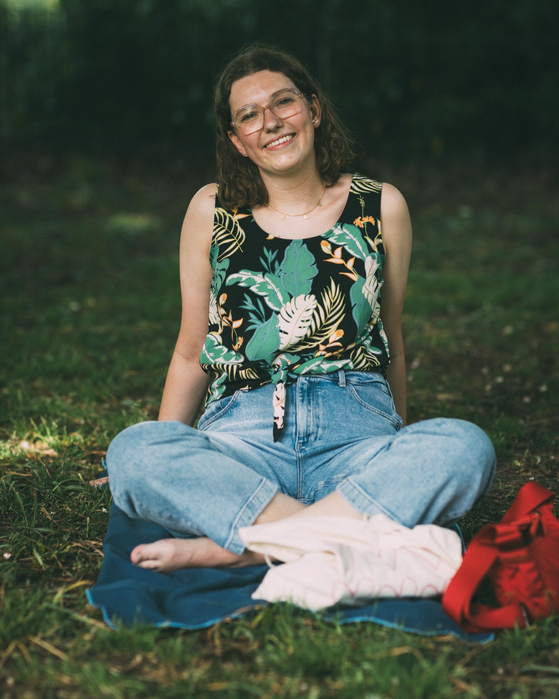 A woman in a leaf-print top sits cross-legged on a blanket in the shade, smiling.