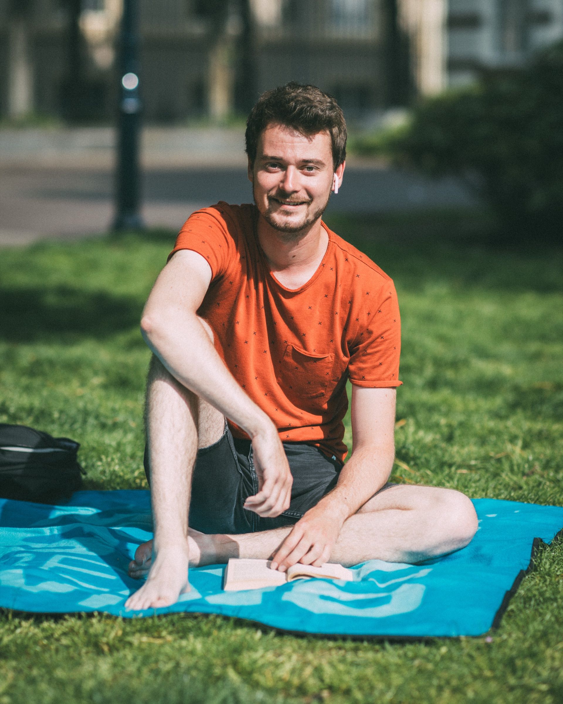 A man in an orange T-shirt sits barefoot on a turquoise towel on the grass.