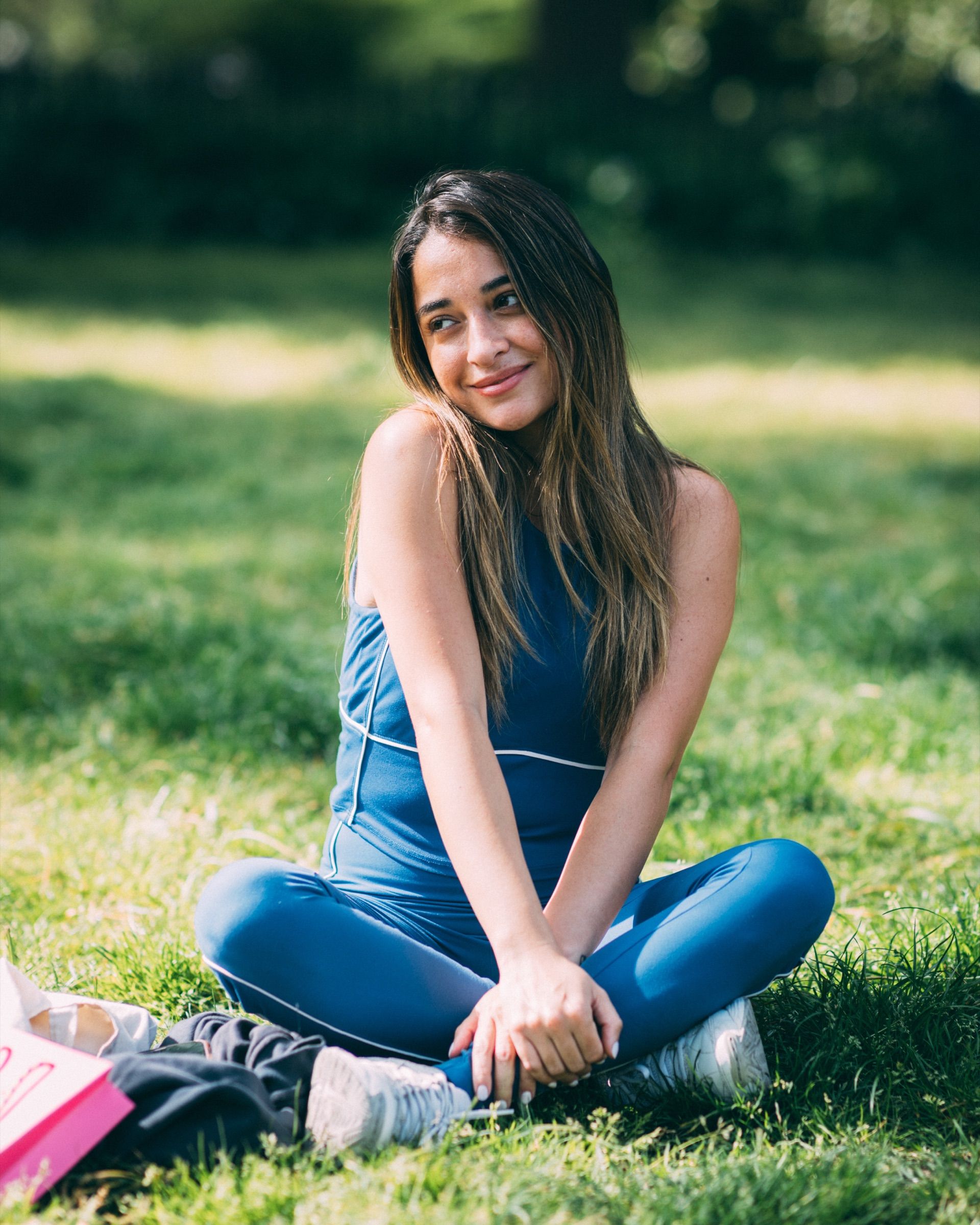 A woman in blue sits cross-legged on the grass and smiles to one side.