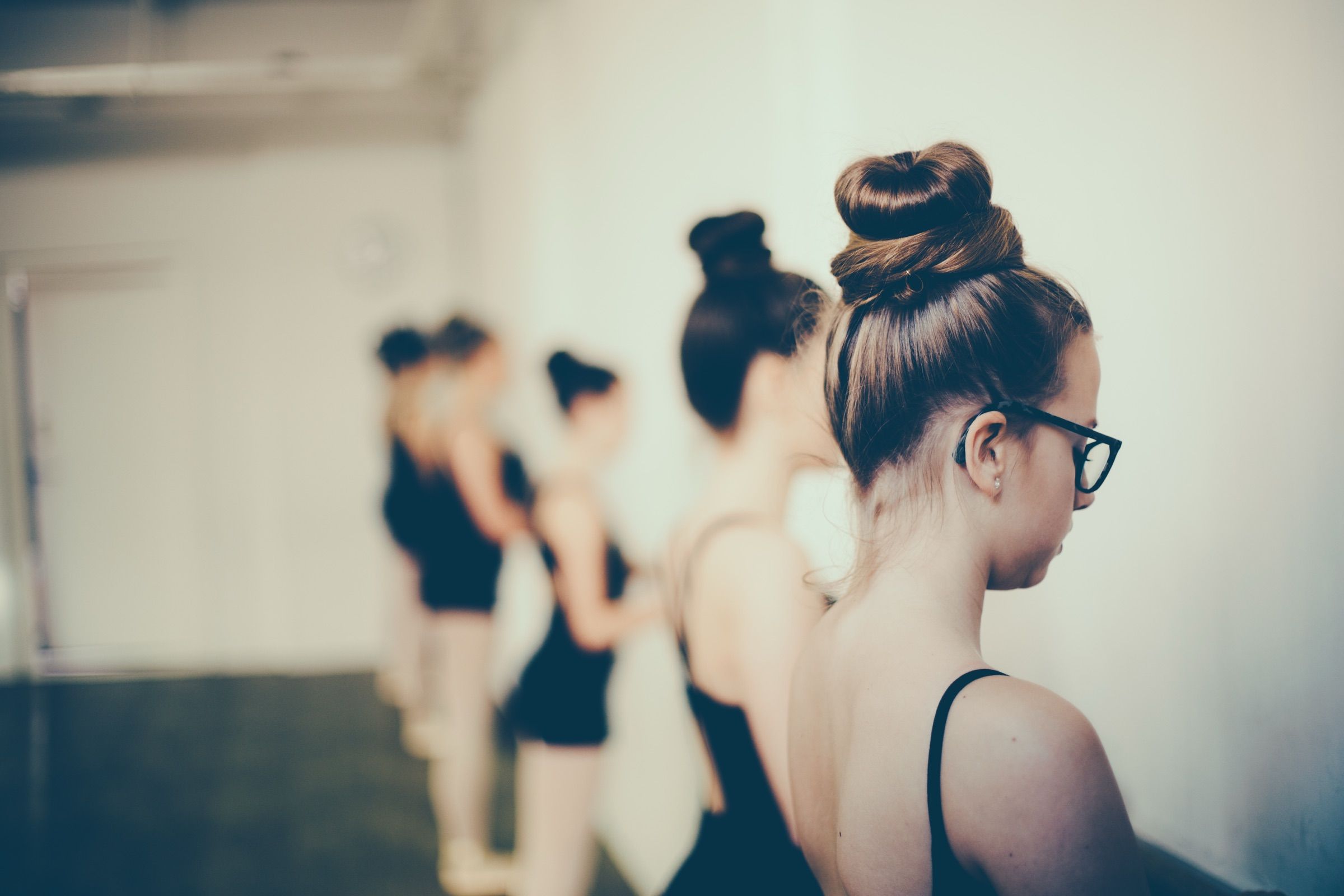 Young dancers in black leotards wait in line at the barre; the nearest, in glasses and a tight bun, looks down.