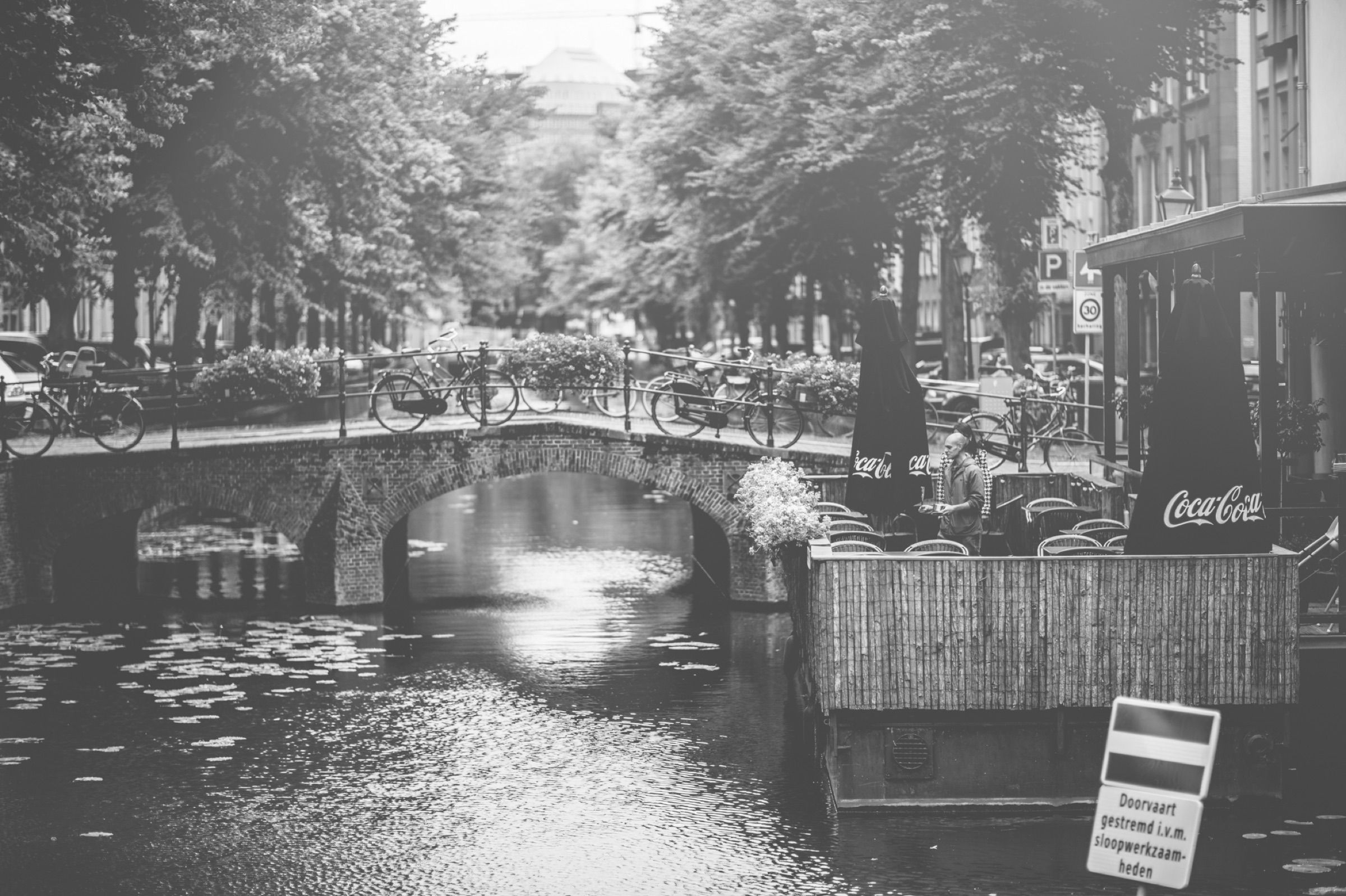 Black and white: a tree-lined canal with a small bridge, the water bright with reflected sky.