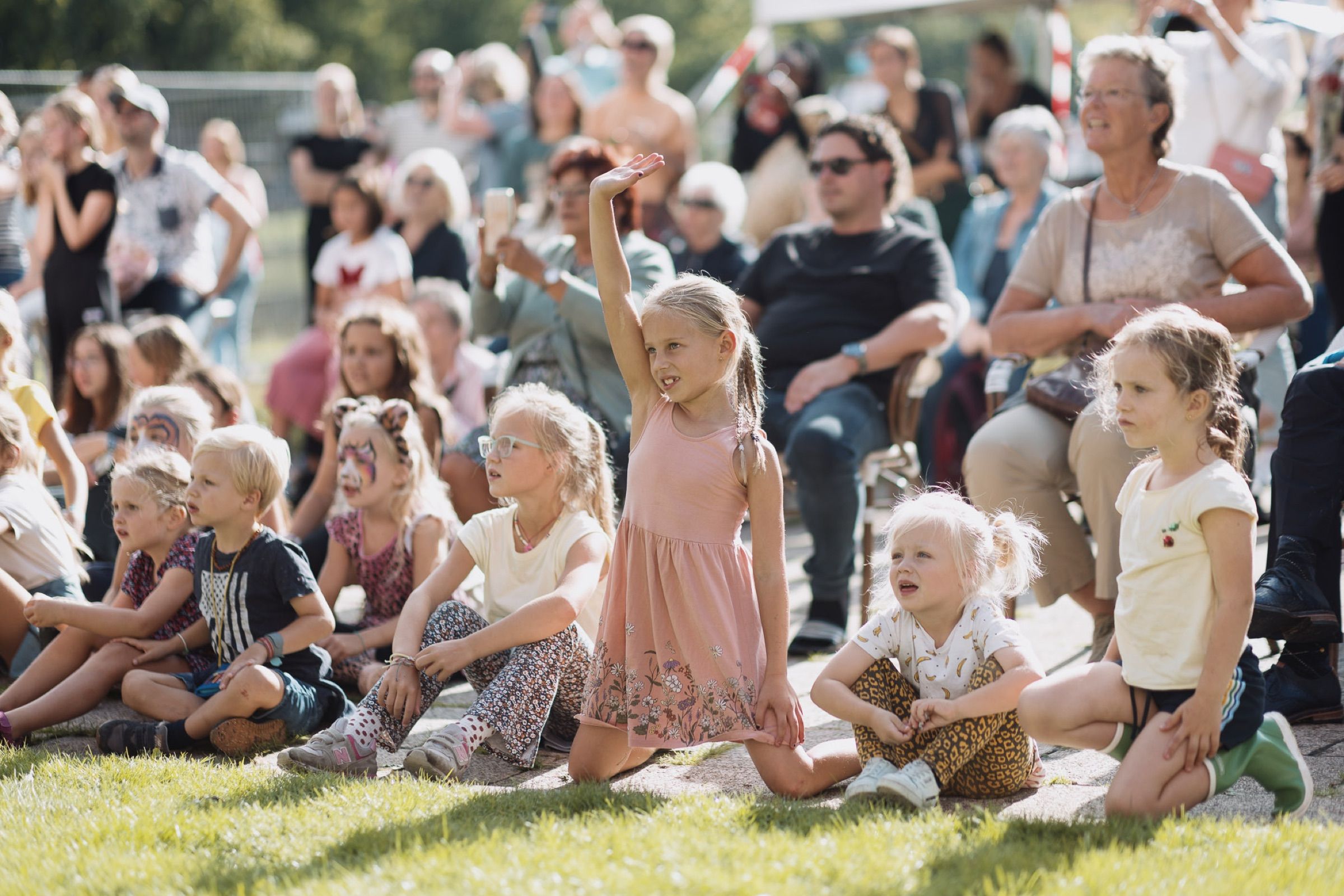 A crowd sits on the grass watching something out of frame; a girl at the front raises her arm.