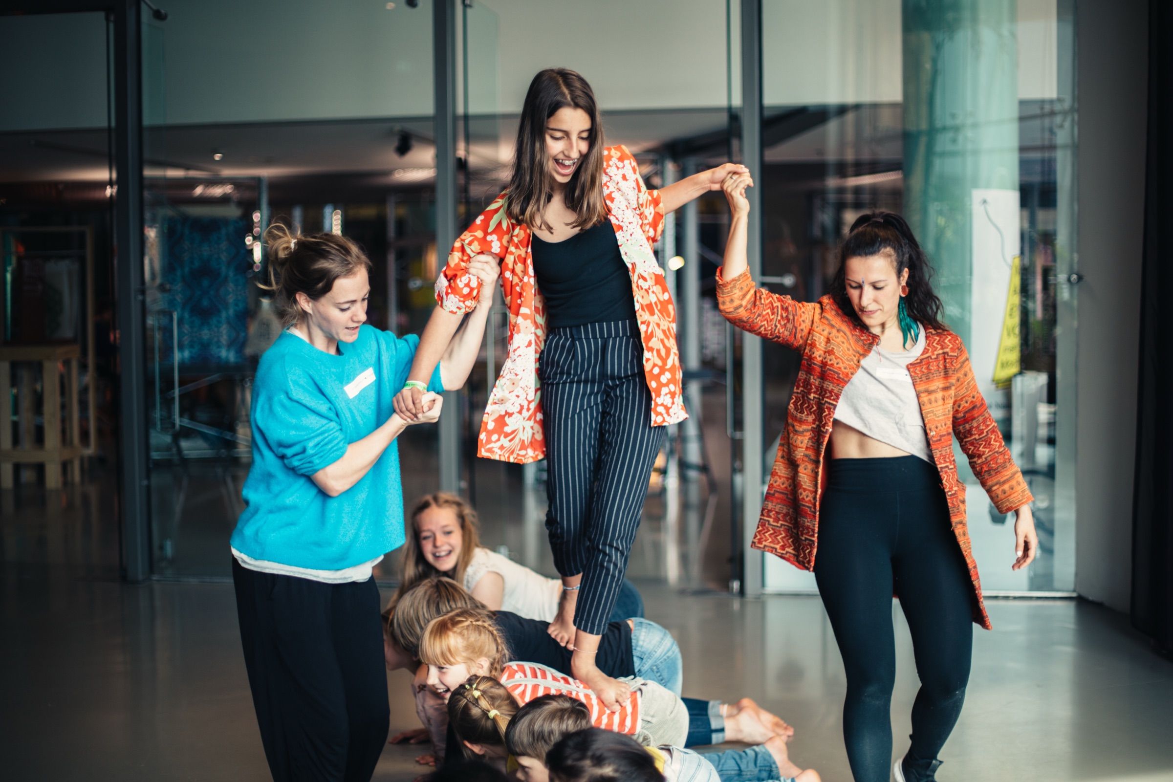 In a bright hall, two women in patterned blouses lead a dance with children, some of them lying on the floor.