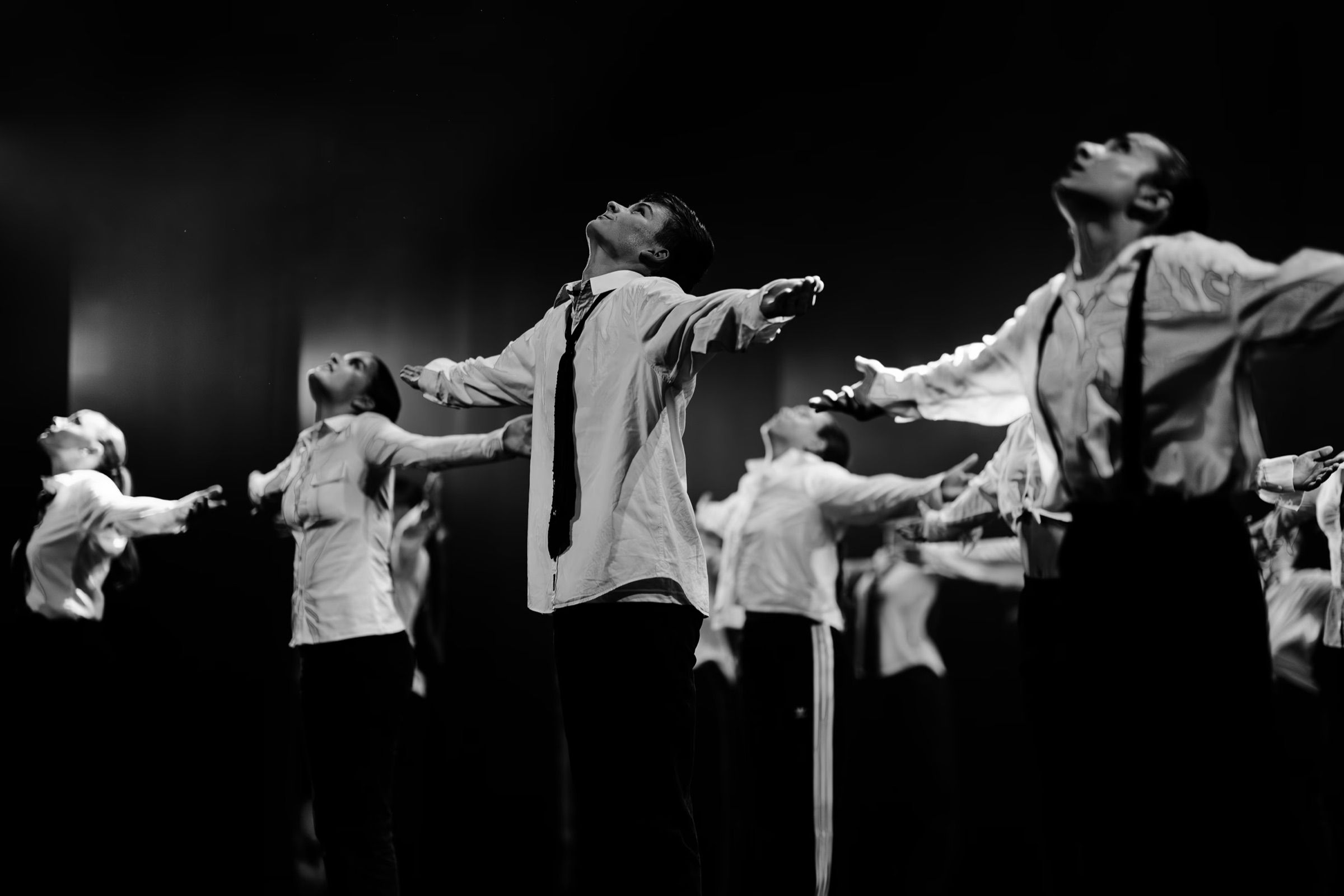 Black and white: a line of dancers in white shirts and braces reaches upward on a dark stage.