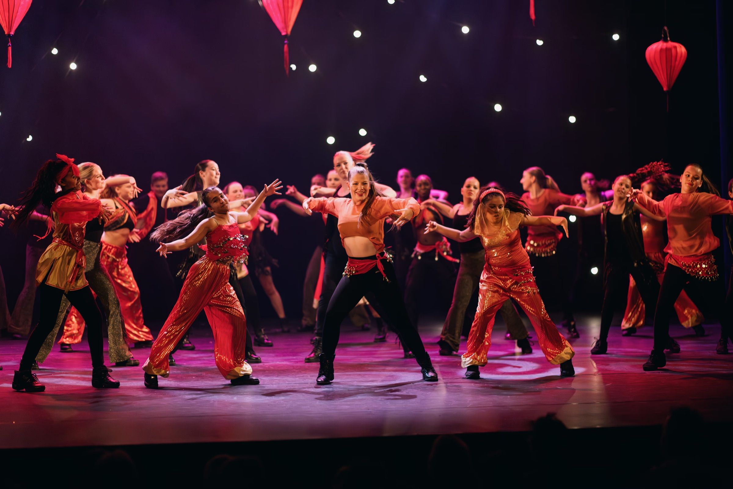 A large group of dancers in red and orange costumes performs under hanging red lanterns and pink stage light.