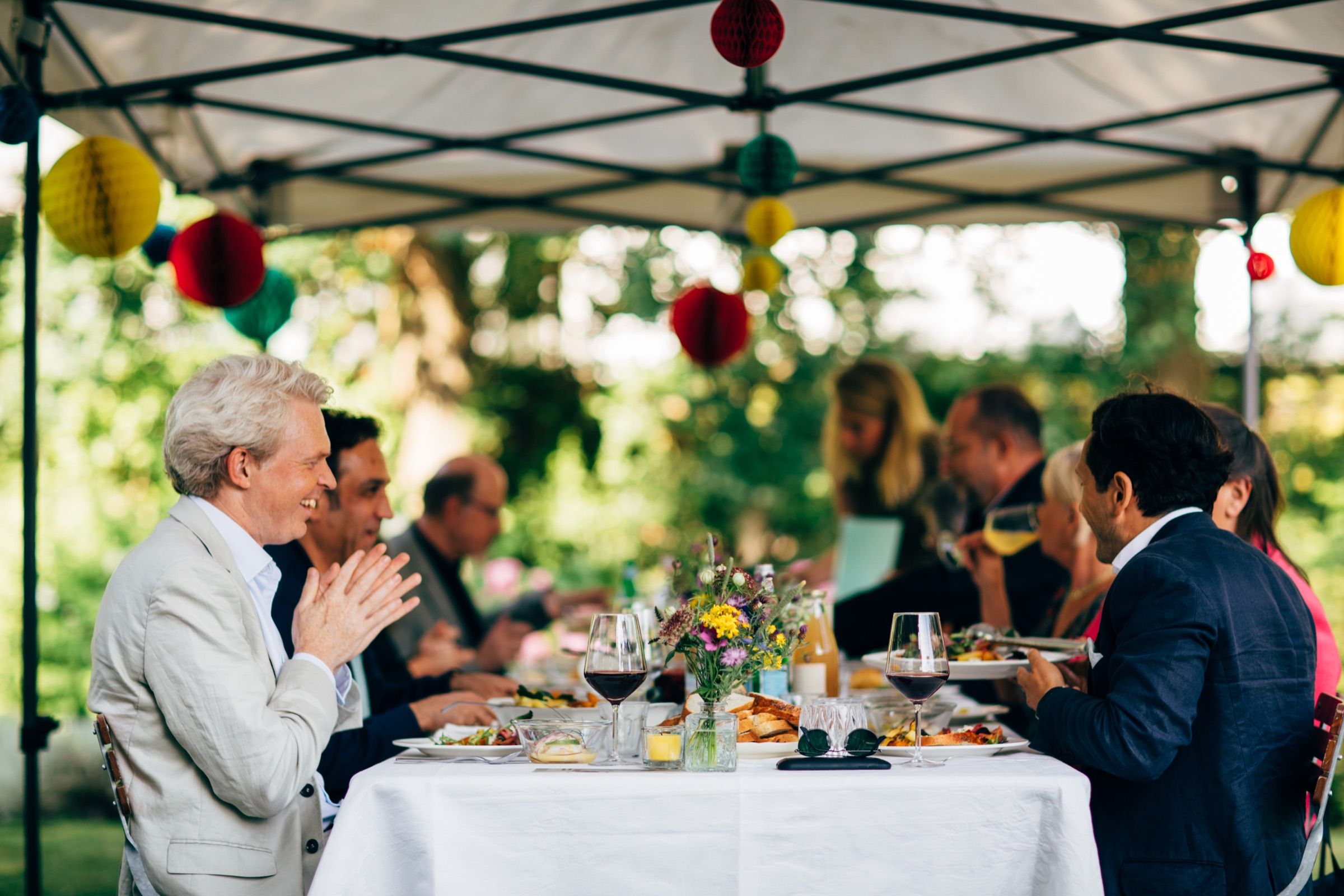 Guests in jackets talk across a long dinner table under a garden marquee hung with paper lanterns.