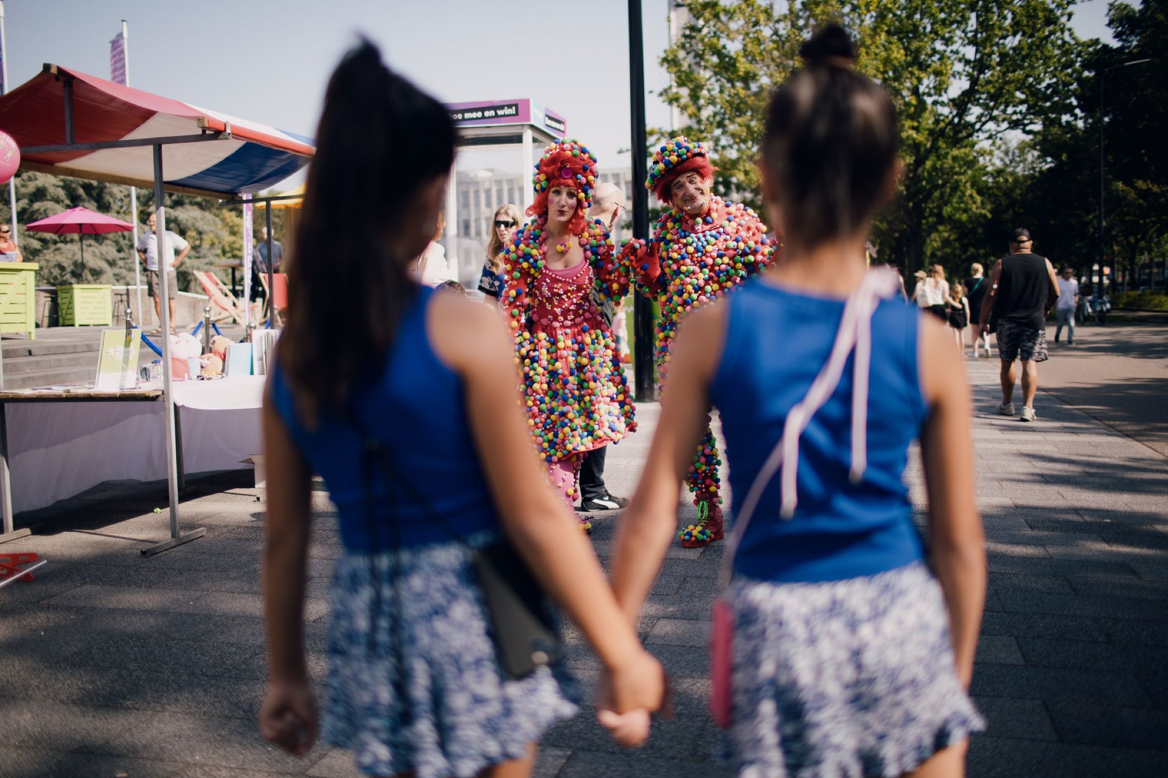 Two girls in matching blue tops and skirts walk hand in hand through a street festival.