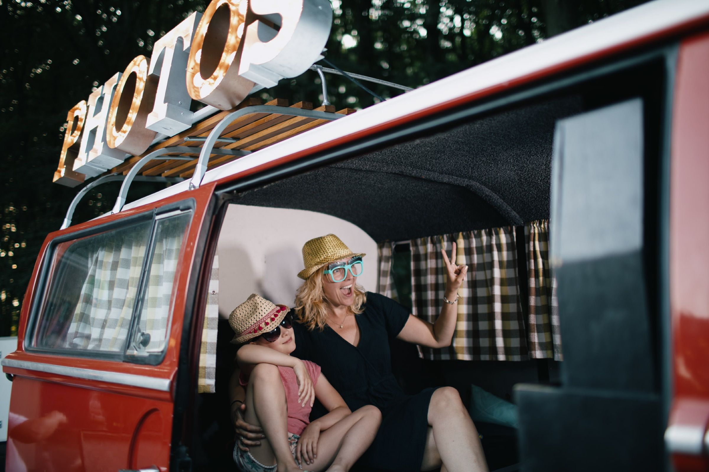 Friends pose inside a red vintage van fitted out as a photo booth, a big PHOTOS sign on its roof.