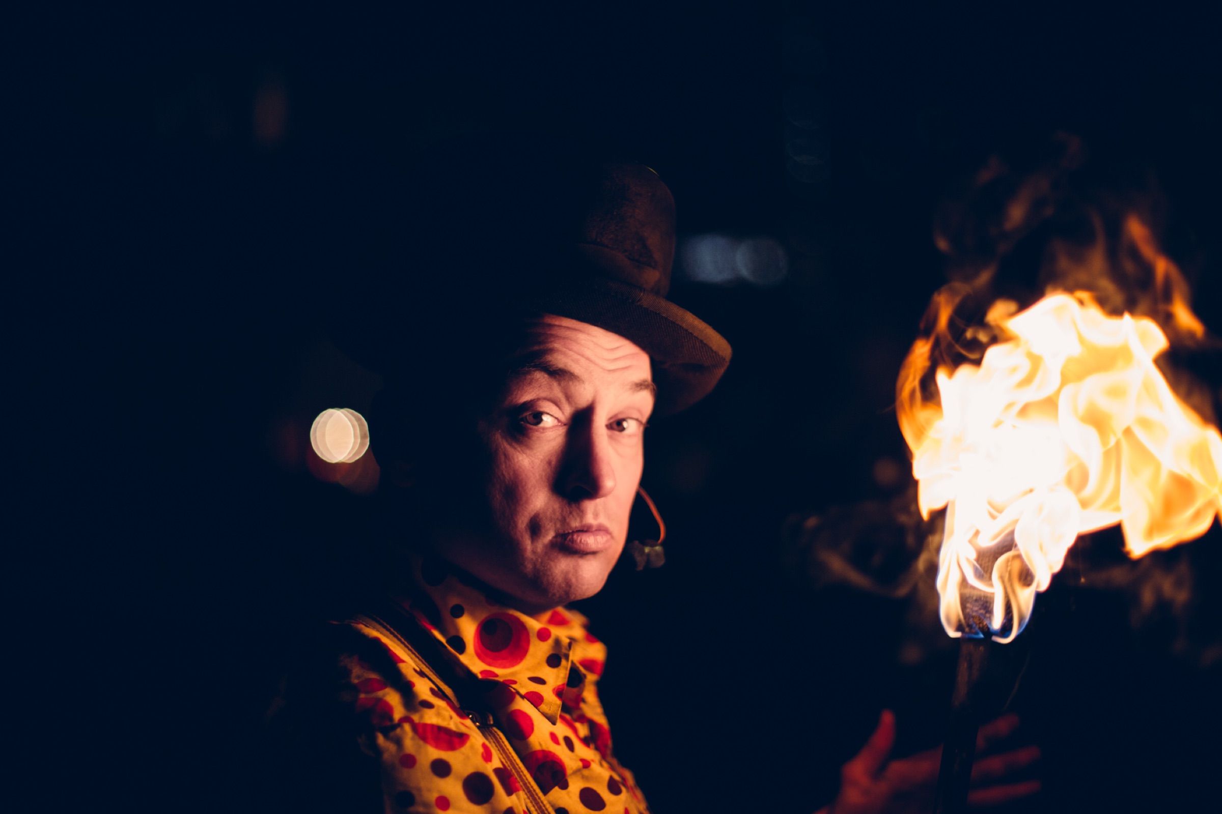 At night, a performer in a hat and spotted shirt looks into the camera, a burst of flame beside him.