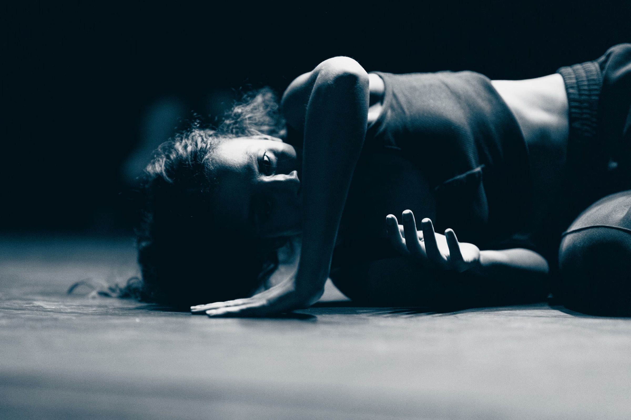 In cold blue light, a dancer arches low over the stage floor, one hand spread on the boards.