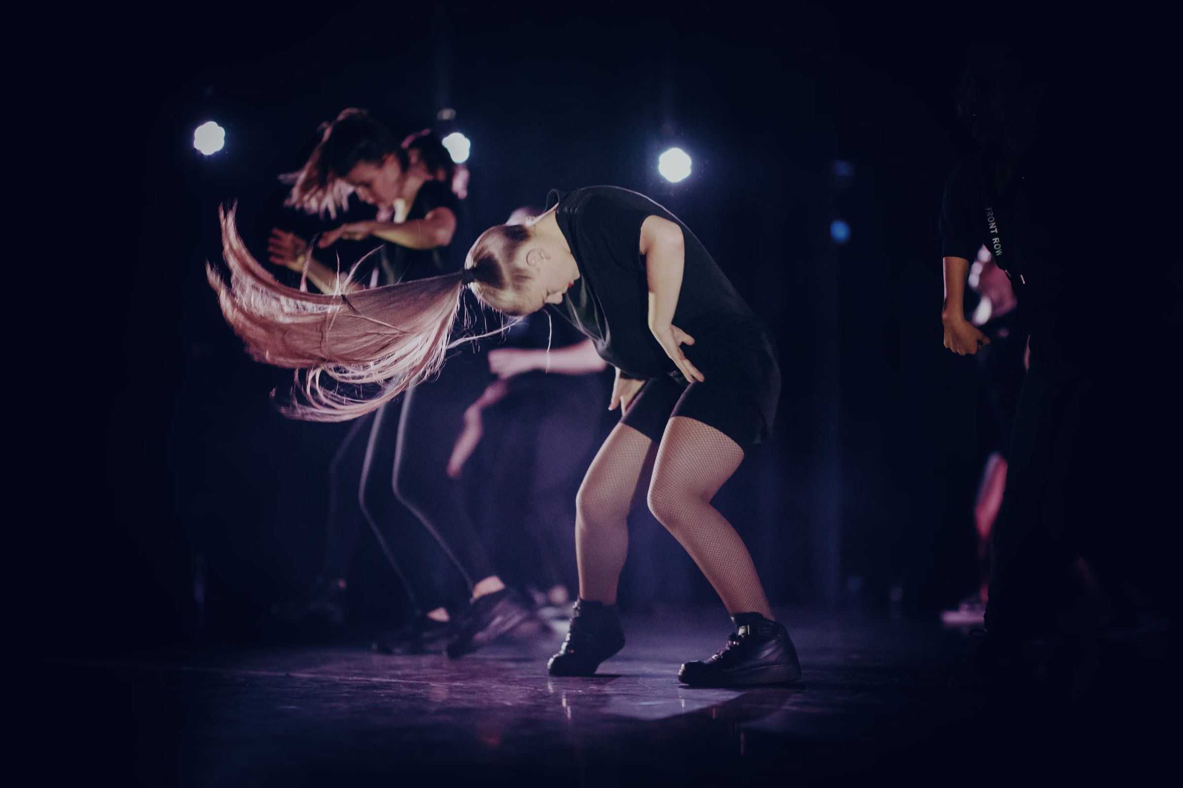 On a dark stage in violet light, a dancer whips her long hair through the air beside her partner.
