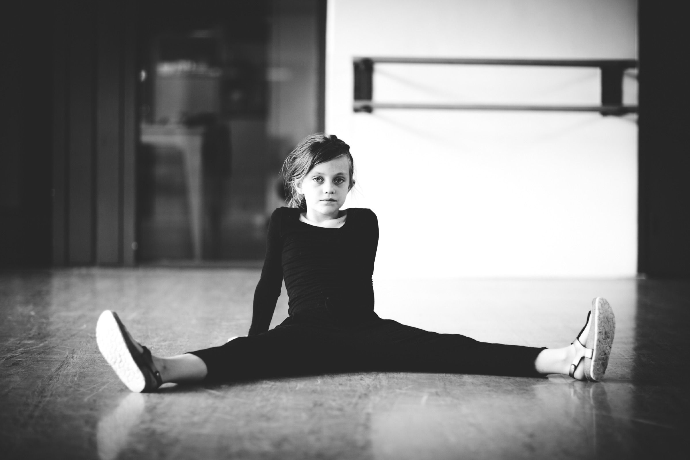 Black and white: the same girl sits in a wide straddle in an empty, bright studio.
