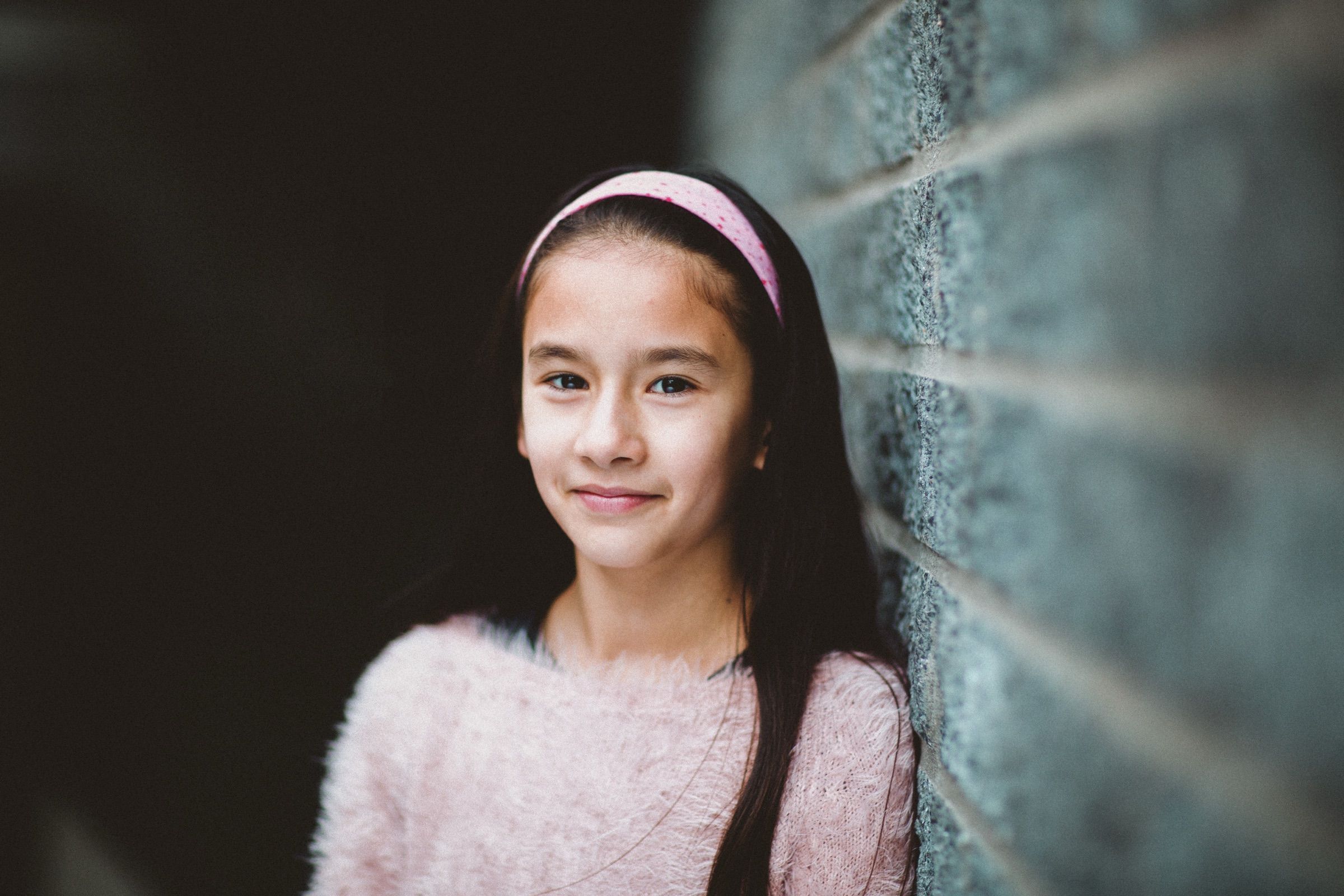 A smiling girl in a pink jumper and hairband leans against a brick wall.