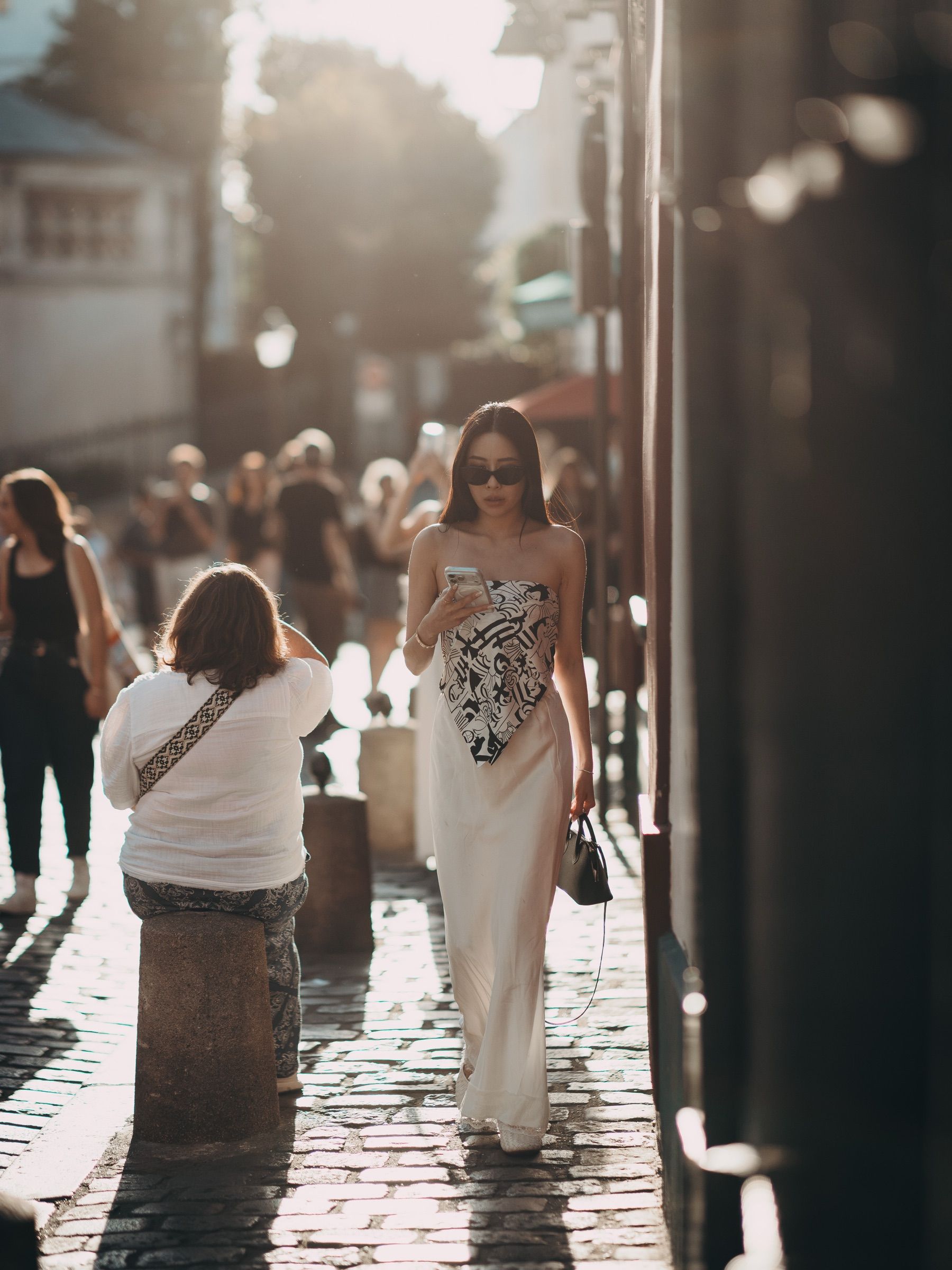 A woman in a white dress walks down a cobbled street, the sun behind her.