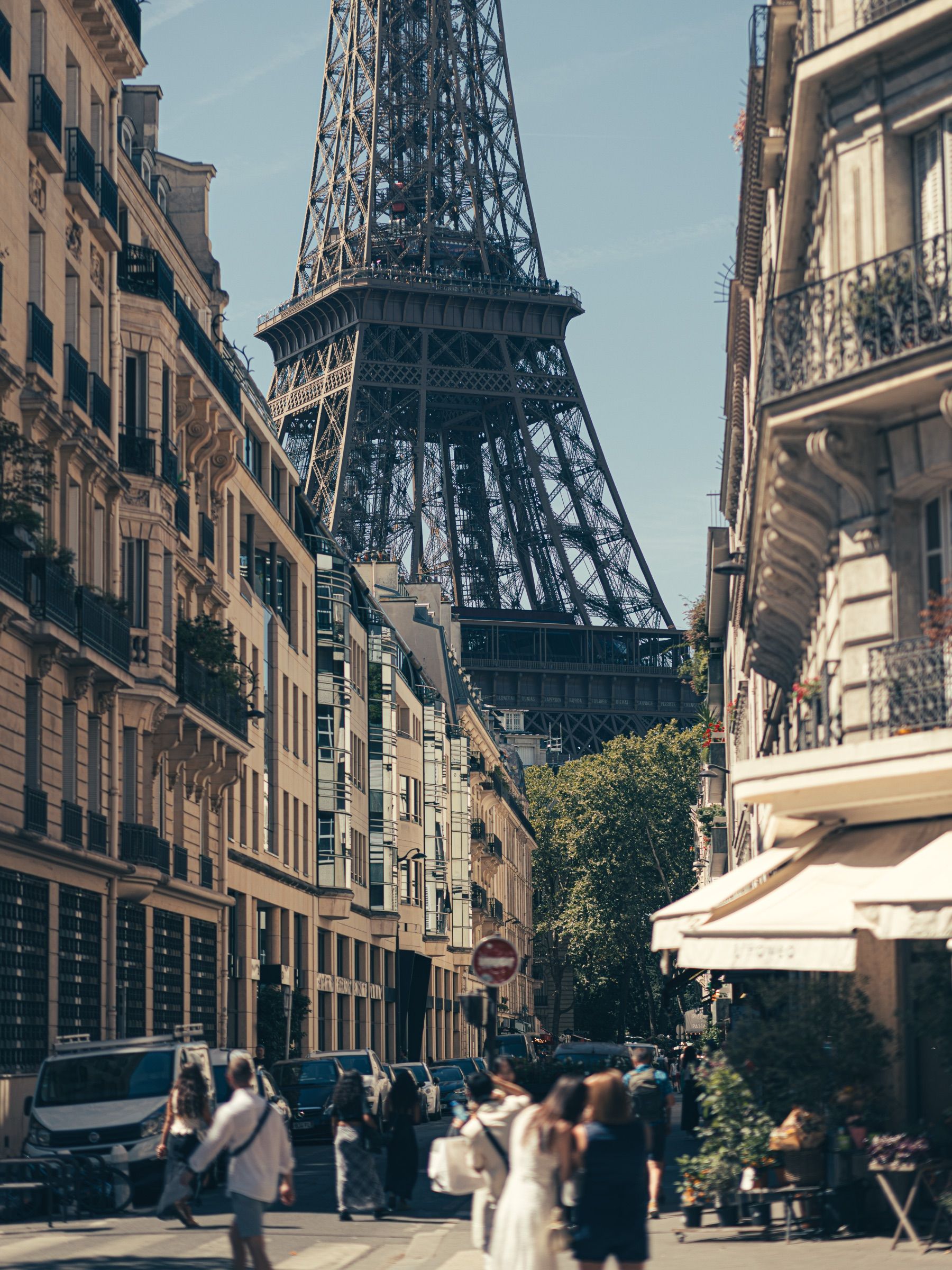 The Eiffel Tower rises at the end of a Paris street of cafés and parked cars.