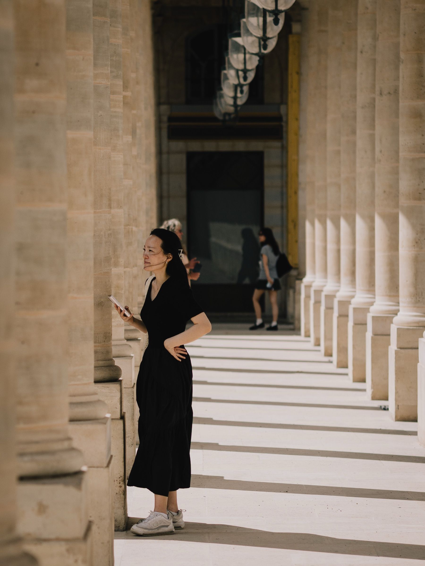 A woman in a black dress stands among the stone columns of a long arcade, light falling across the floor.