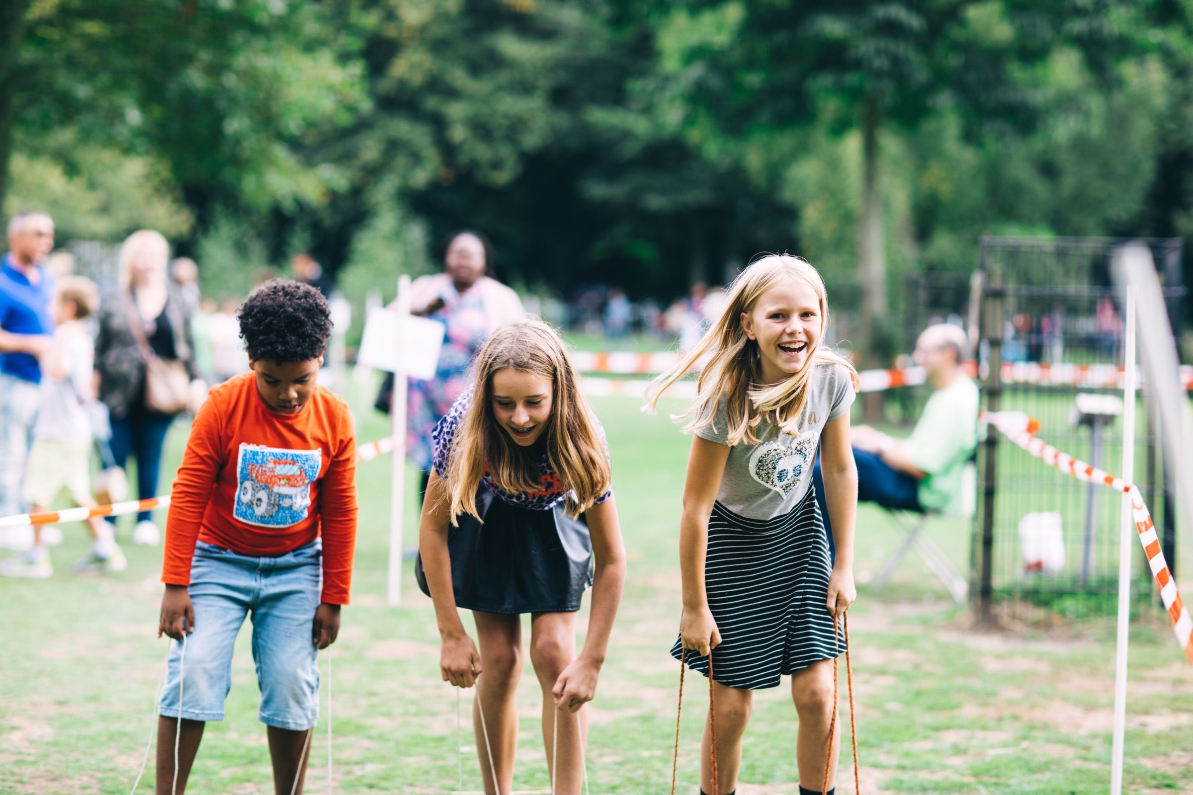 Three children bend forward, laughing, at a game on a park lawn during a festival.