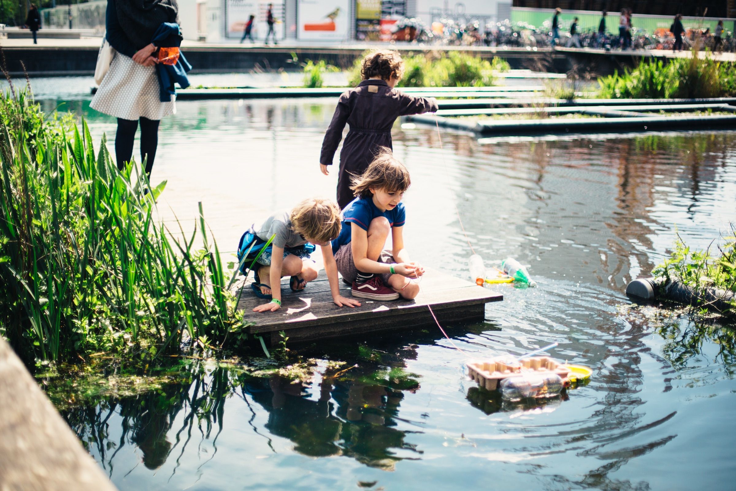 Children kneel on a small raft on a city pond and reach into the water while people pass on the bank.