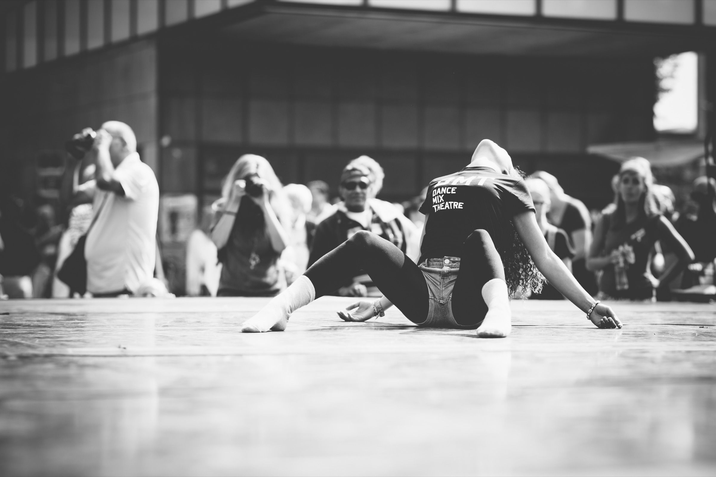 Black and white: dancers stretch and rest on a rehearsal floor while others sit watching.