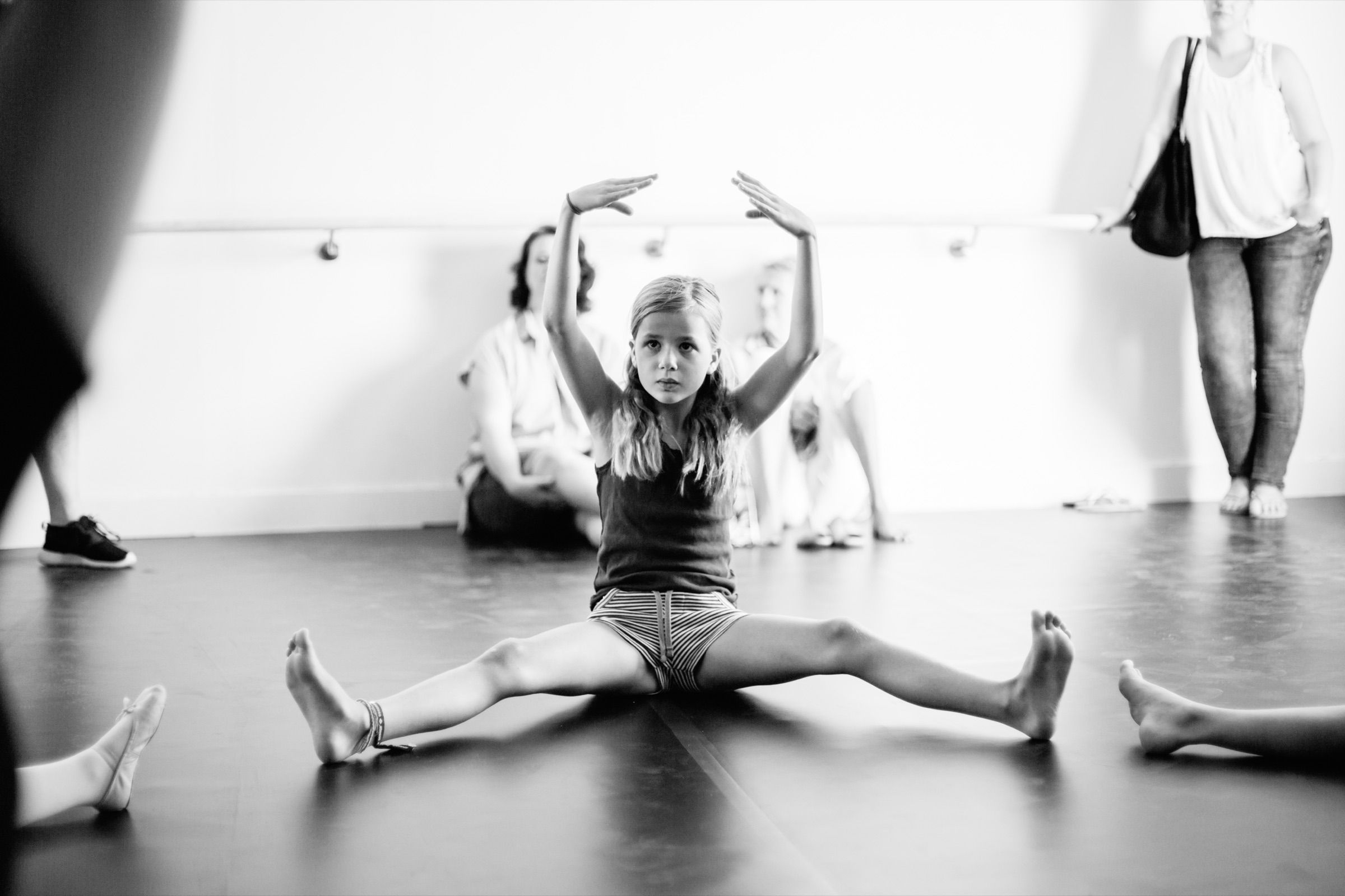Black and white: a girl sits in a split on the studio floor, arms raised, other dancers resting along the wall.