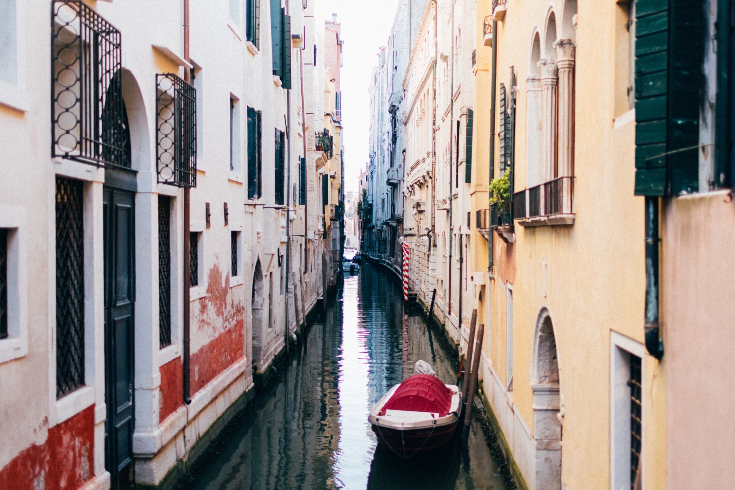 A narrow Venetian canal between ochre and white houses, a red boat moored below.