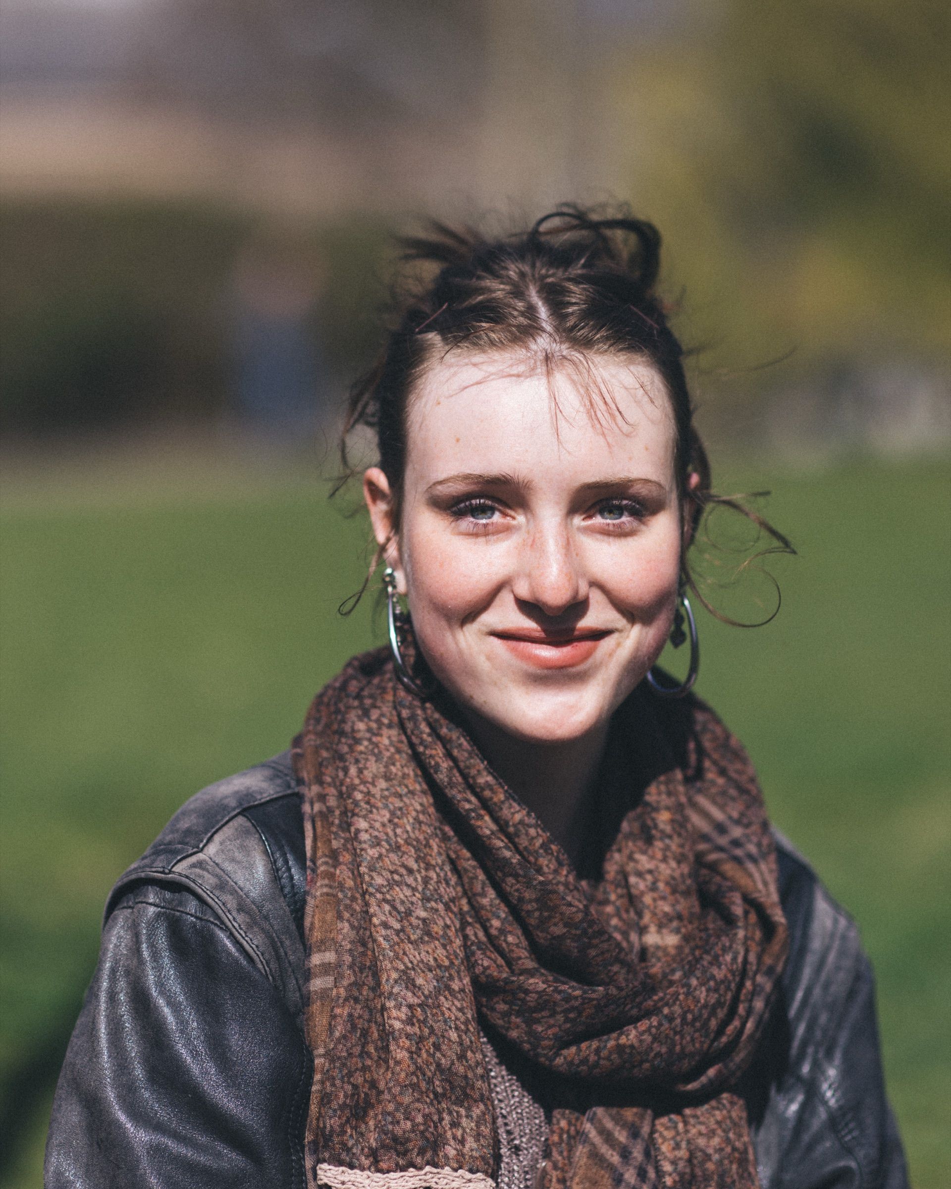 Een jonge vrouw met een bruine sjaal en krullend haar lacht in een zonnig park.