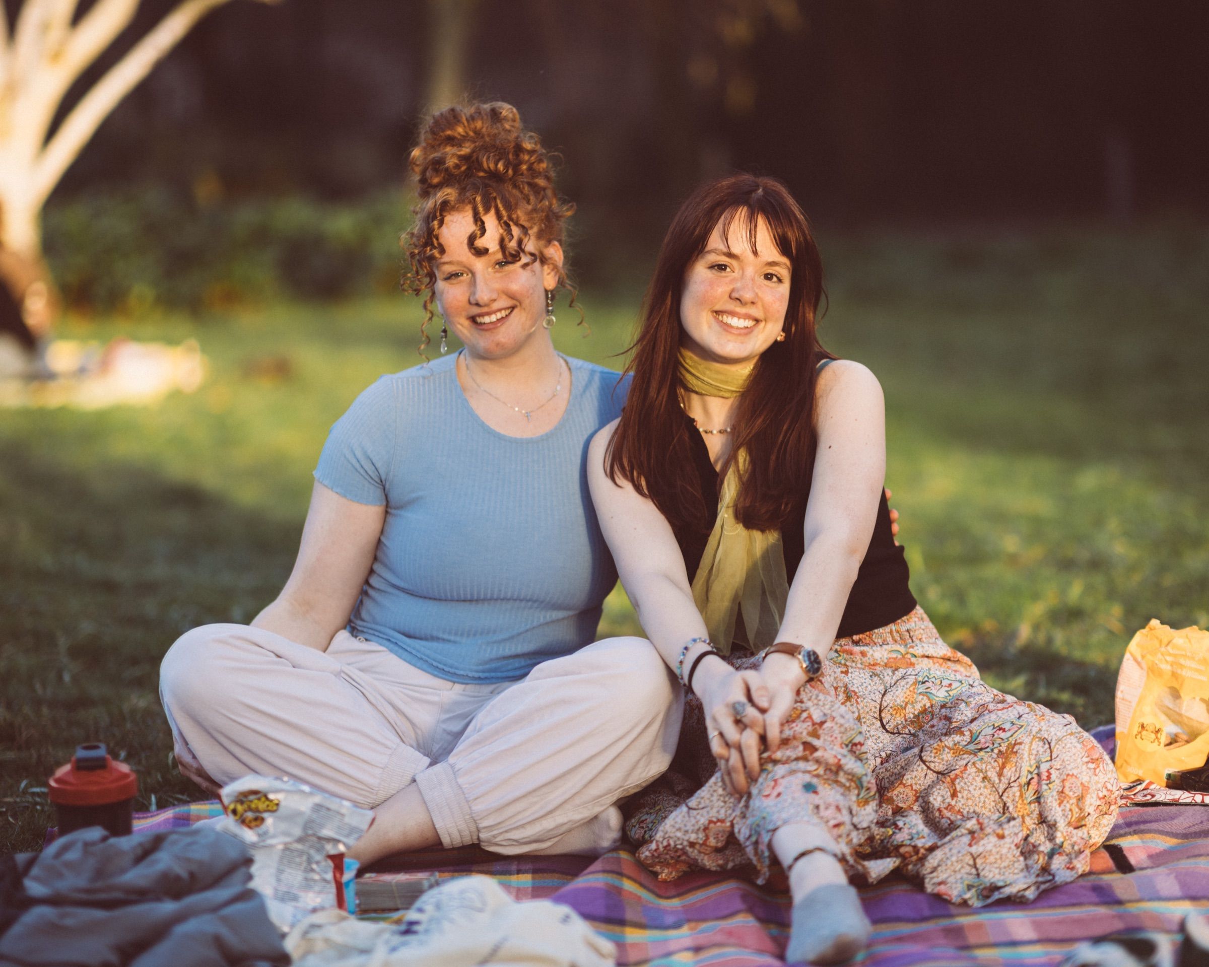 Twee jonge vrouwen zitten dicht tegen elkaar op een picknickkleed in laat middaglicht.