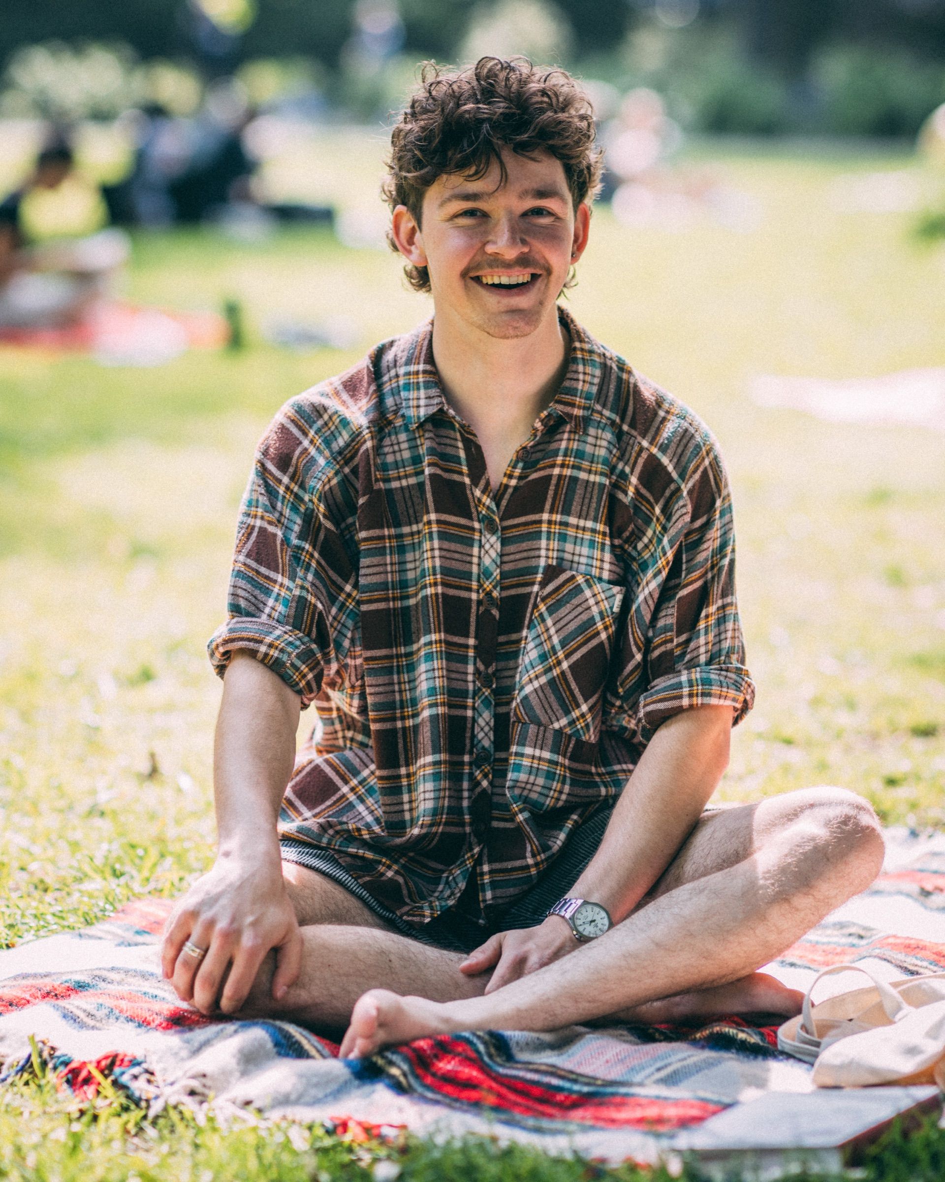 Een jonge man in een geruit overhemd zit in kleermakerszit op een kleed in het gras en lacht.