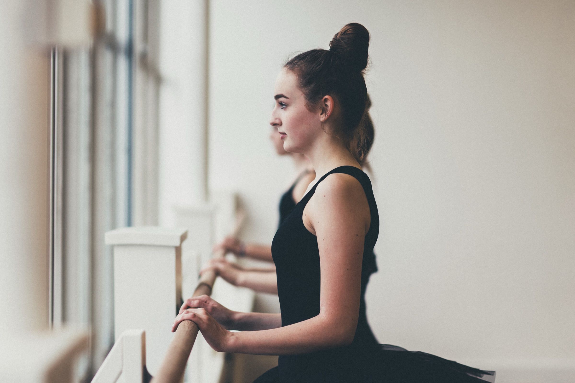 Een jonge danseres met haar haar in een knot houdt de barre vast bij een licht studioraam.