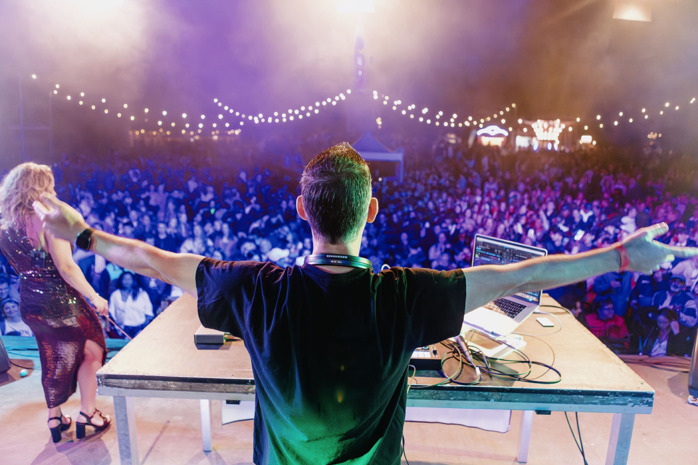 Van achteren gezien spreidt een dj zijn armen naar een festivalpubliek onder paars licht en lichtslingers.