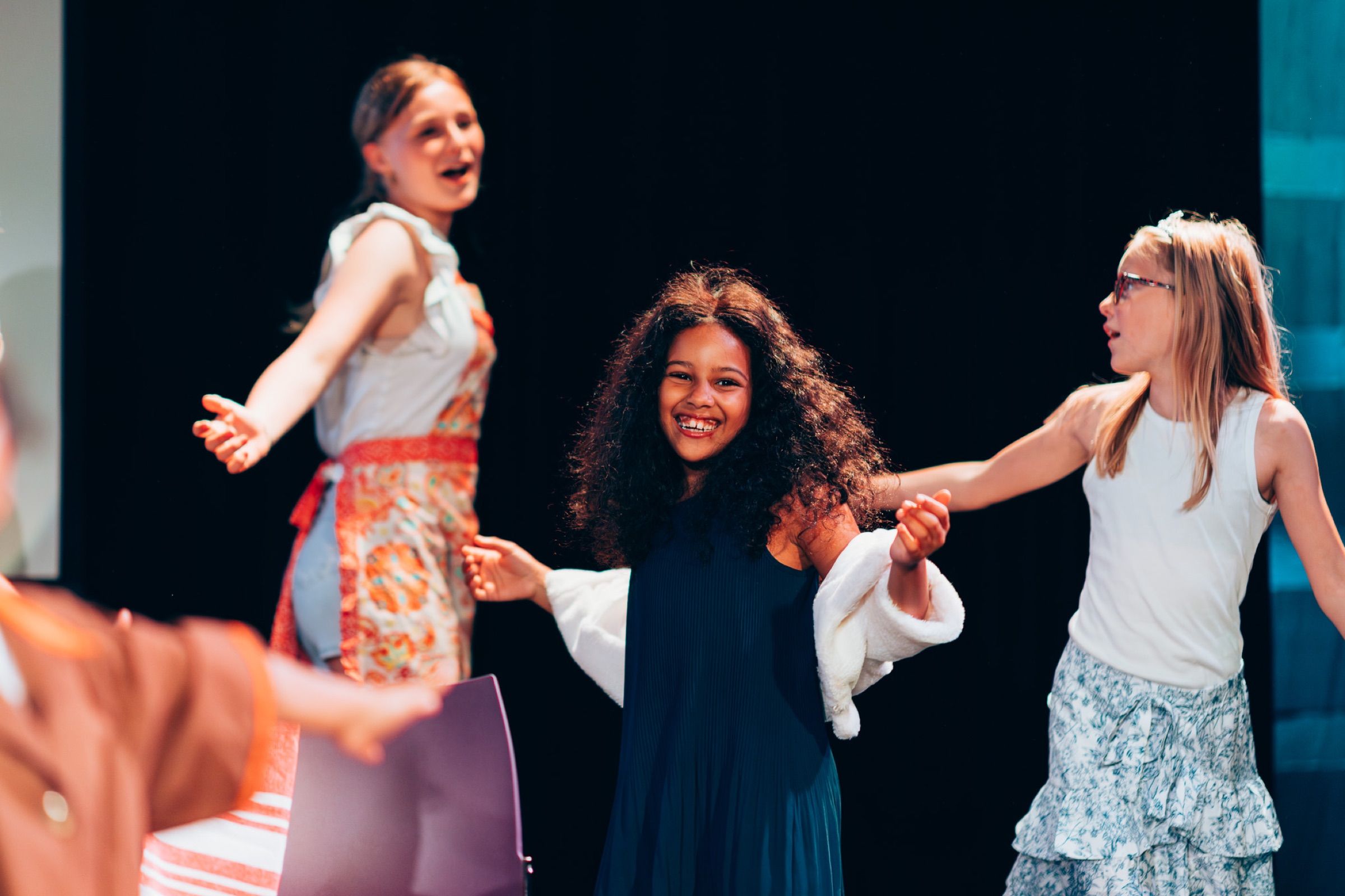 Drie meisjes in kostuum dansen hand in hand op een musicalpodium, zingend, tegen zwart.