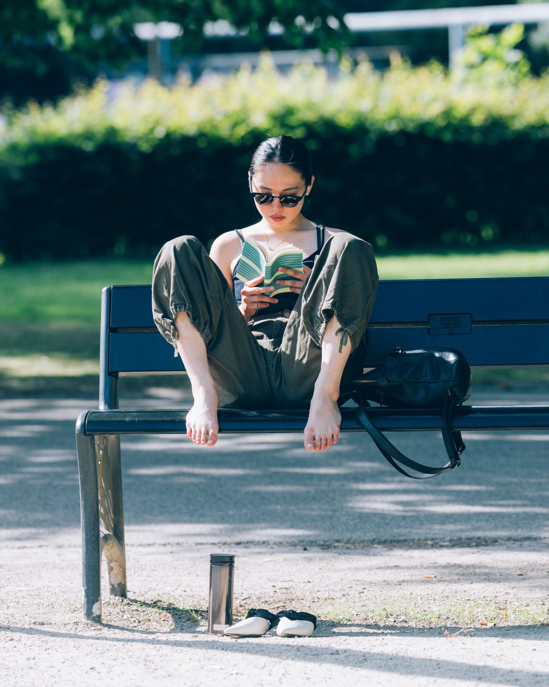 Een vrouw met zonnebril leest op een parkbank, haar schoenen uit op het grind.